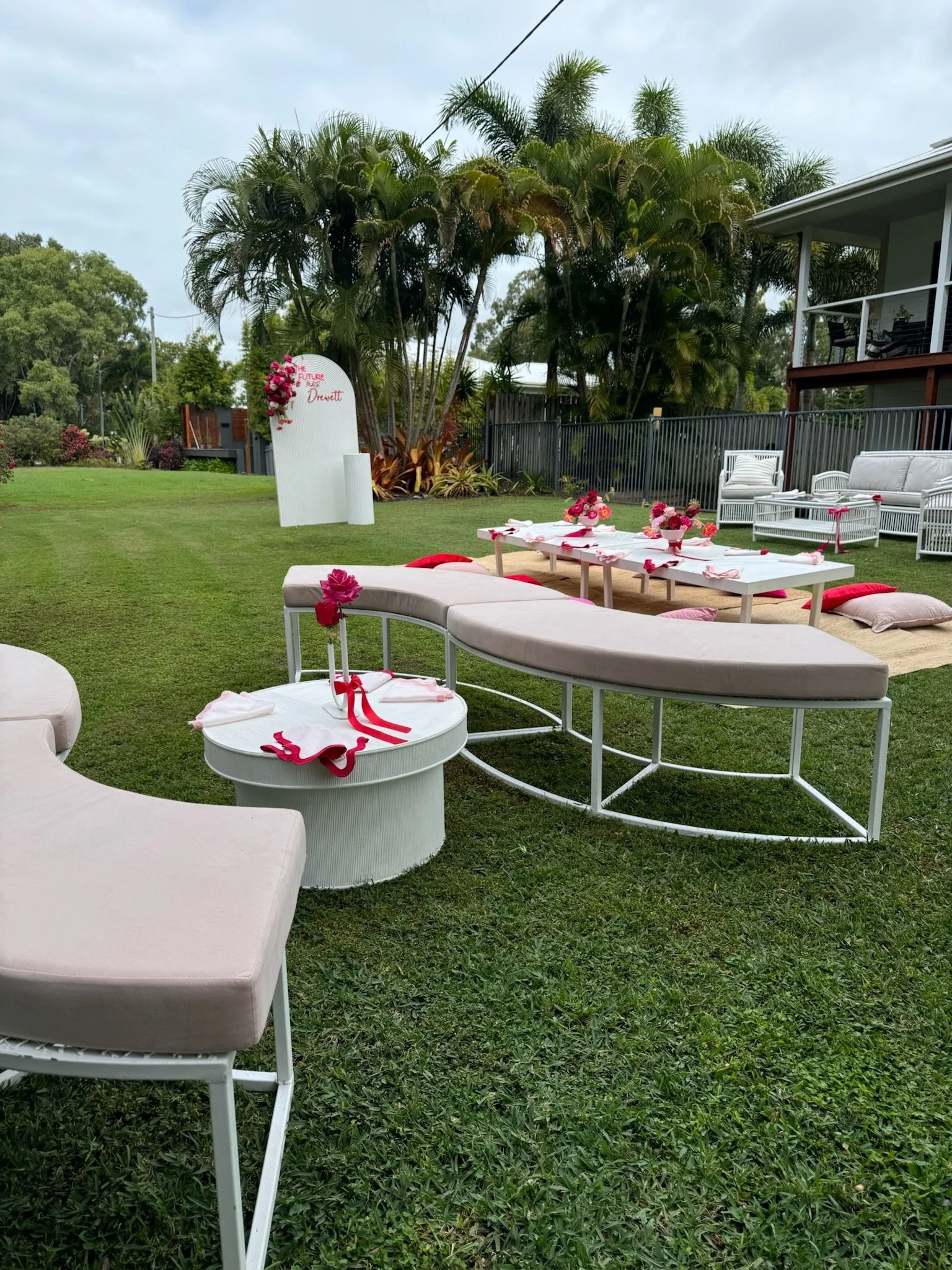 Outdoor setup with white and pink furniture, flowers, and a grassy yard, decorated for a bridal or bridal shower event.
