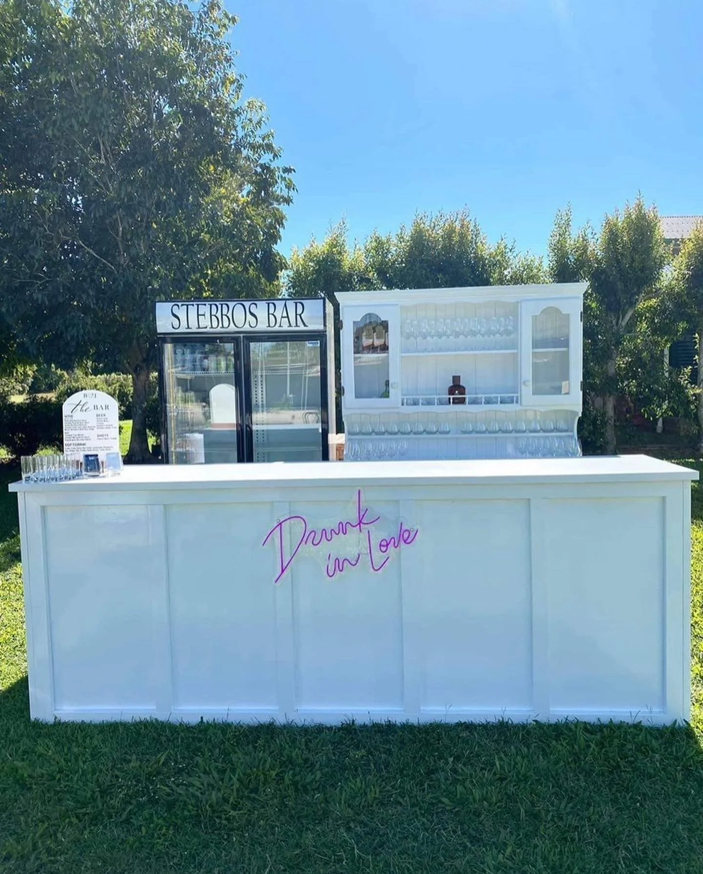 White outdoor bar with neon pink sign reading 'Drunk in Love', set up in a grassy area with trees, featuring a small fridge labeled 'Stebbos Bar' and glassware display.