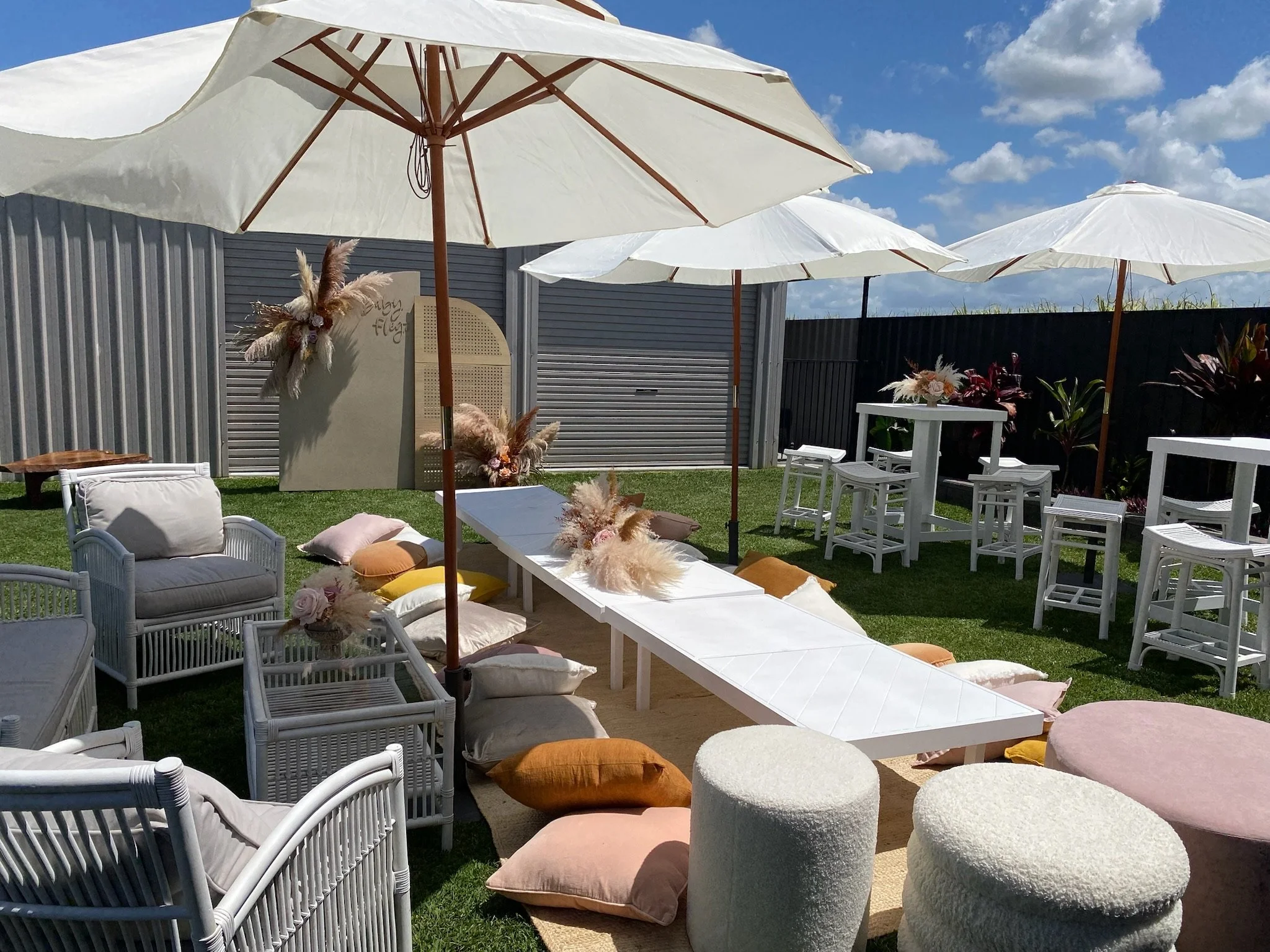 Outdoor patio setup with white umbrellas, pastel cushions, and white furniture on a sunny day with a blue sky and clouds.