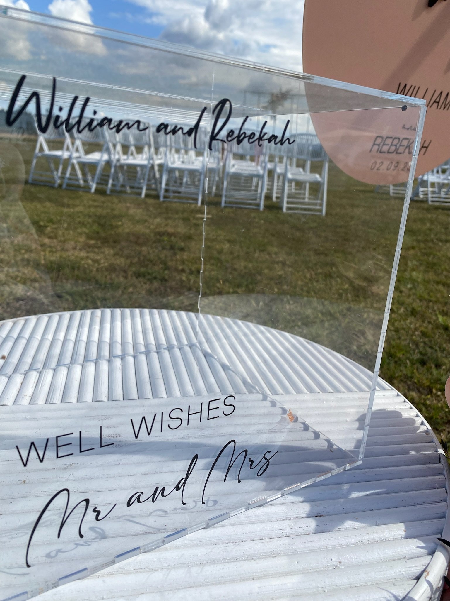 A clear acrylic wedding sign on a white, ribbed table reads "WILLIAM and REBEKAH" in black cursive. In the background, there are white chairs set up outdoors on a grassy area with a partly cloudy sky overhead.