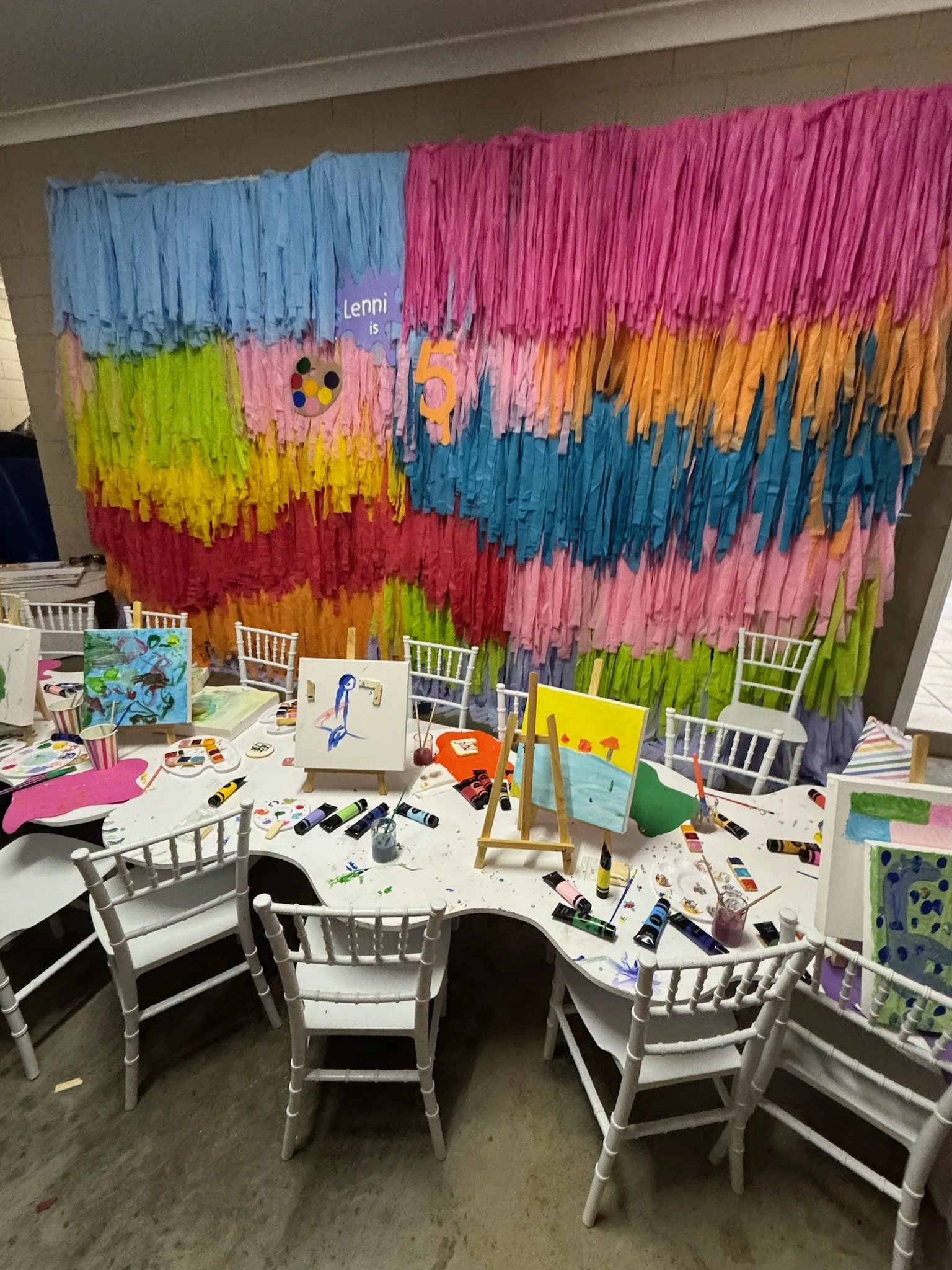A colorful birthday party setup with a decorated backdrop made of multicolored tissue paper streamers hanging on a wall, with a table covered with children's paintings and painting supplies such as paints, brushes, and cups. White chairs surround the
