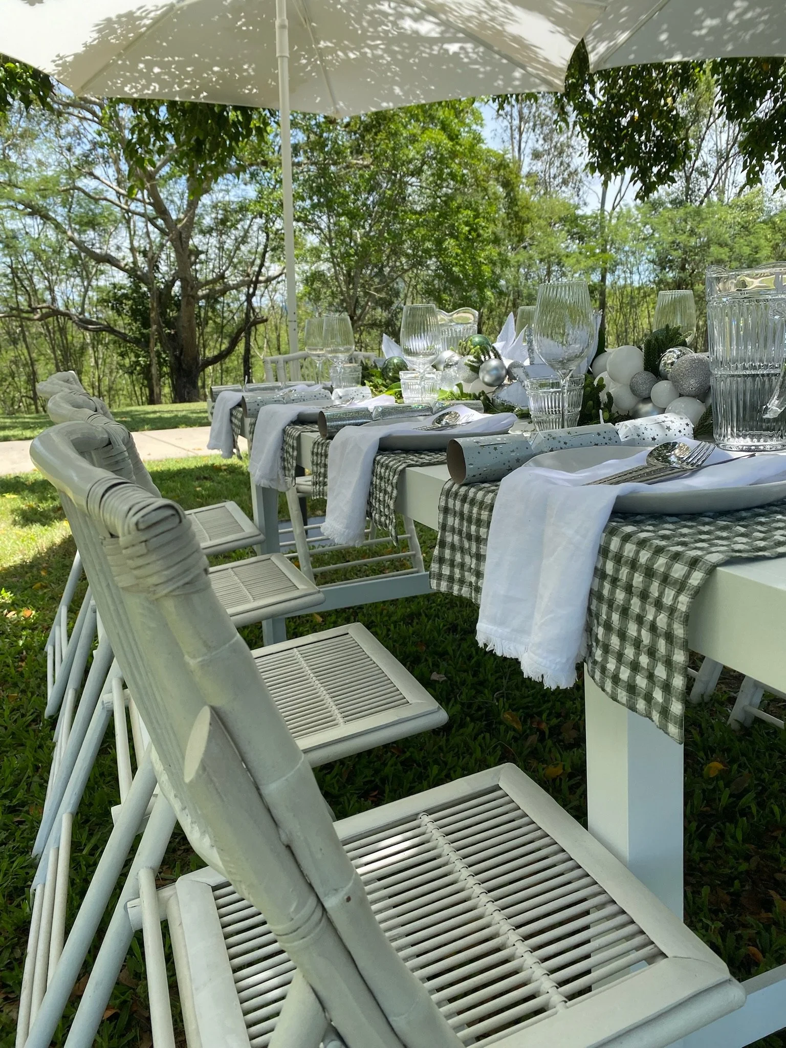 Outdoor dining table set with glassware, plates, silverware, napkins, and decorative ornaments under an umbrella, surrounded by trees.