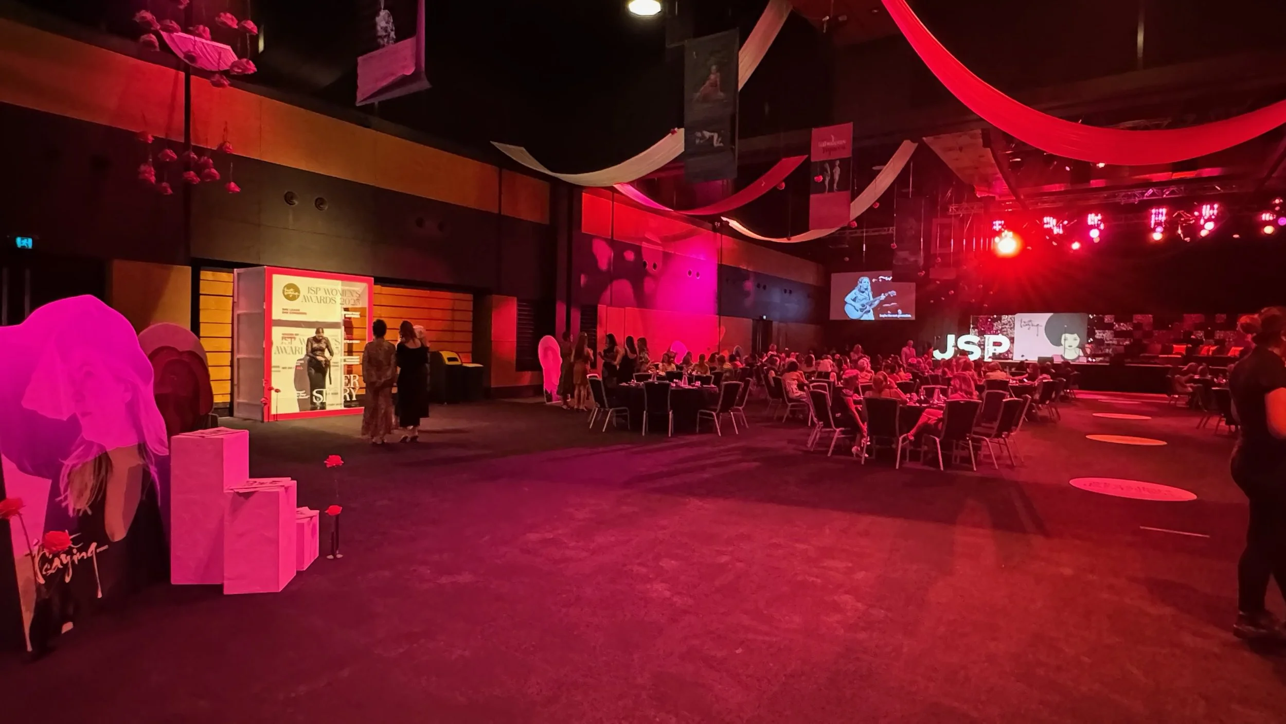 Indoor event venue decorated with pink and red drapes, with a stage displaying the 'JSP' logo, tables with chairs, and a few people mingling near a photo backdrop.