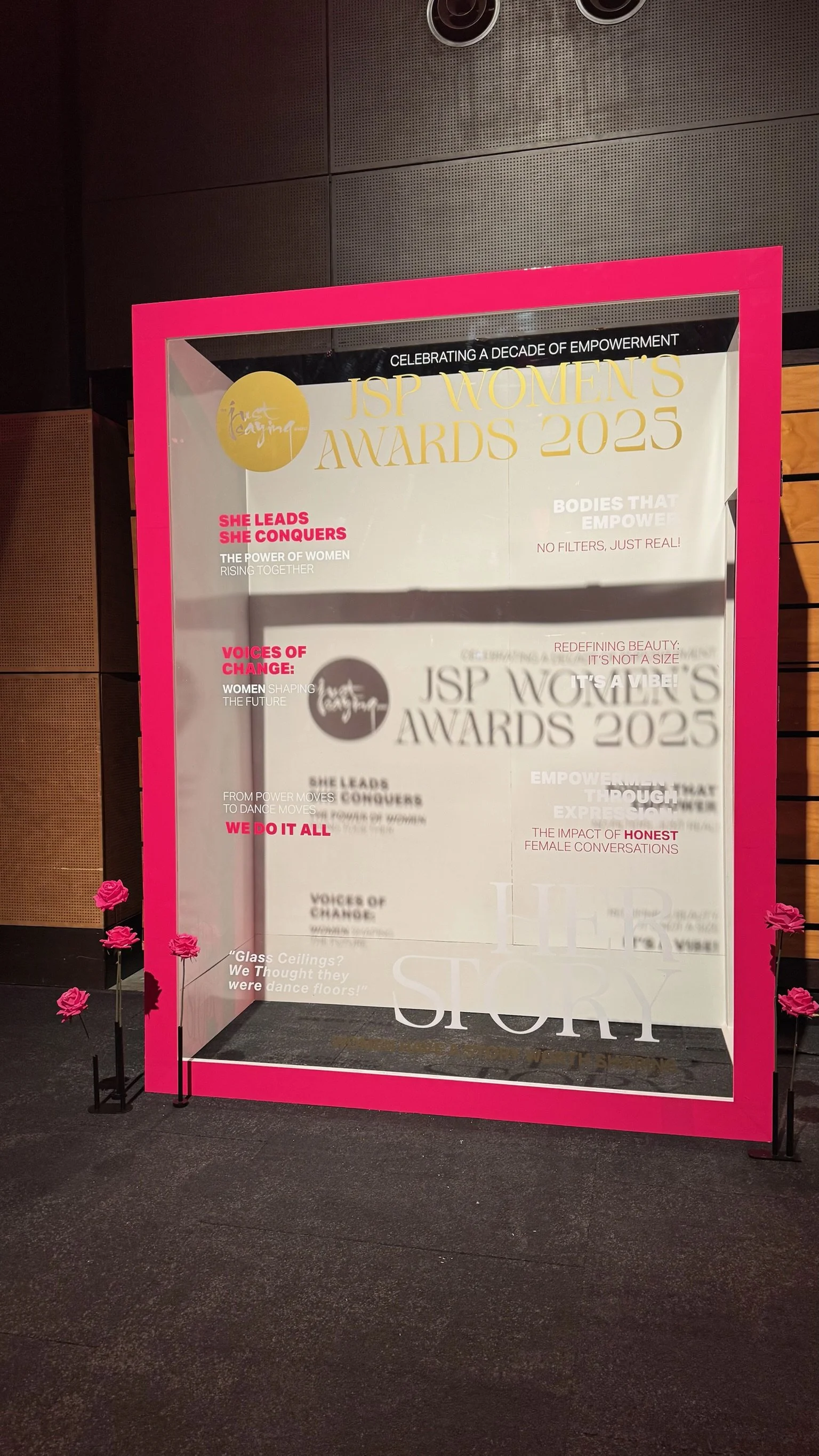 A glass display case at the JSP Women's Awards event with pink border and pink roses at the base, containing a poster about the 2023 awards celebrating female empowerment, voices of change, and honest conversations.