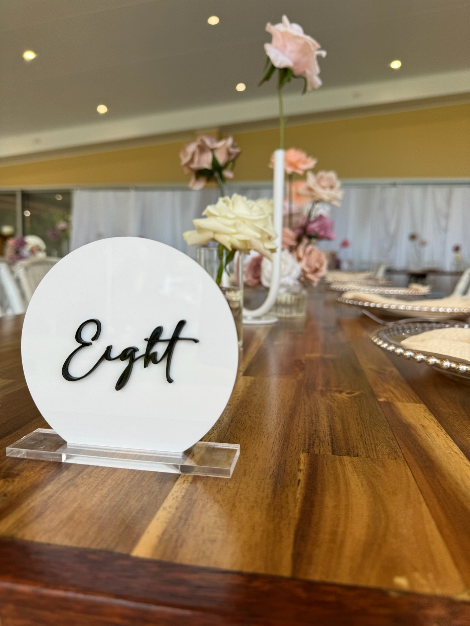 Table number sign displaying 'Eight' in black cursive font on a white circular background, set on a clear acrylic stand, with a centerpiece of pastel-colored flowers in a vase on a wooden table.