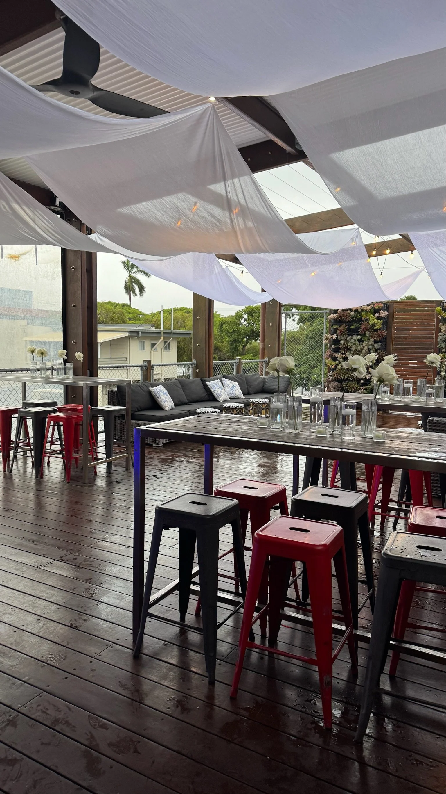Indoor outdoor patio with wooden decking, black and red industrial-style stools, a black sectional sofa with pillows, tables with white flowers in glass vases, white fabric draped ceiling, and string lights, with trees and houses visible outside.