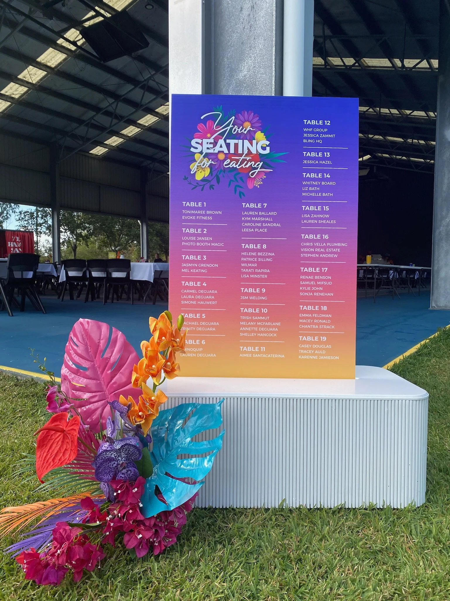 Sign with colorful floral design displaying a seating arrangement for an event, listing table numbers and names of guests or groups, positioned outside under a large covered area.