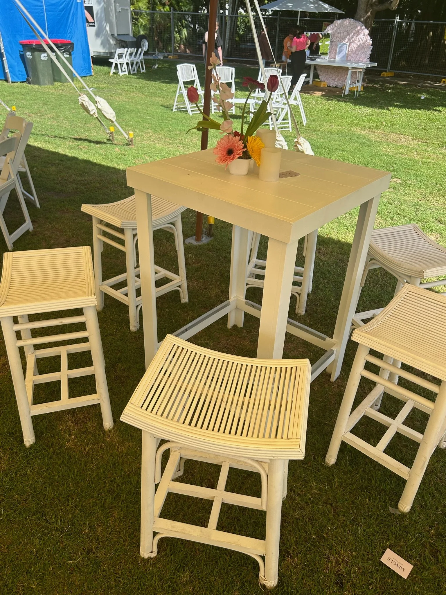 White outdoor table with flowers and candles, surrounded by white chairs and high stools, set on a grassy area at an outdoor event.