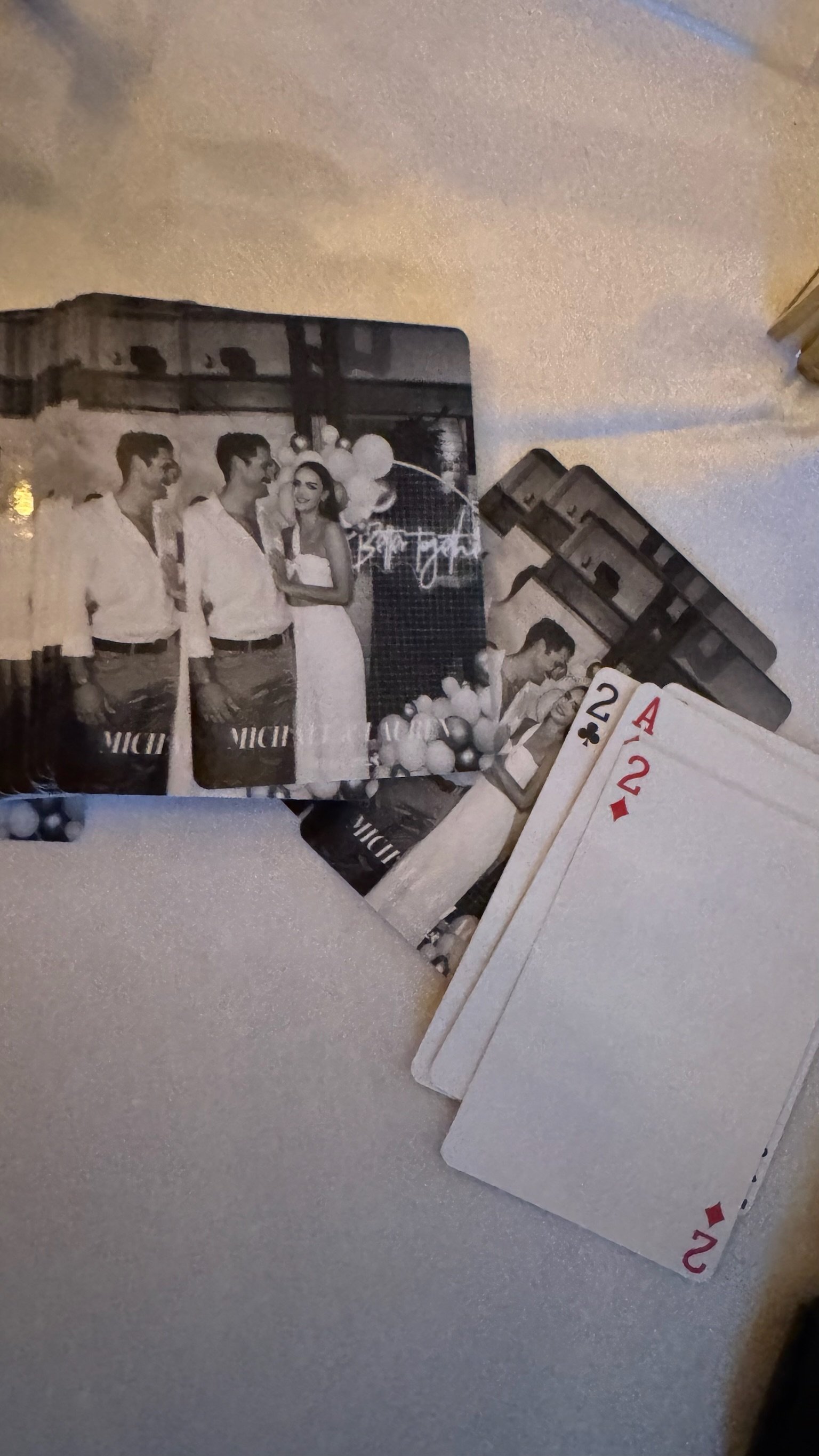 A black and white photograph of four people, three men and one woman, smiling and standing close together at a celebration, with balloons and decorations in the background. There are also playing cards in front of the photo.