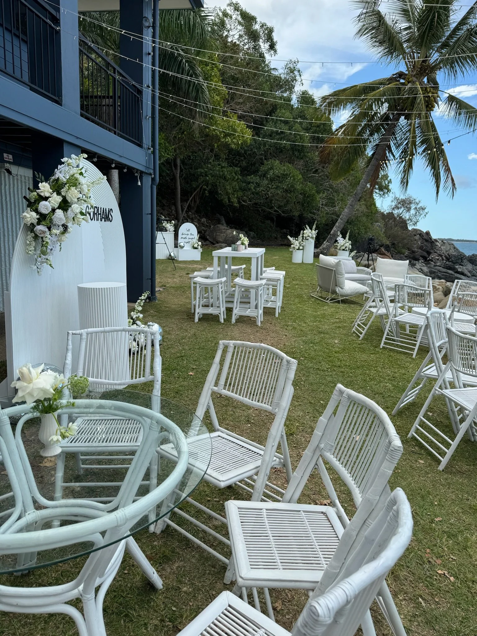 Outdoor wedding setup with white chairs, tables, and floral decorations near the beach, under a palm tree.