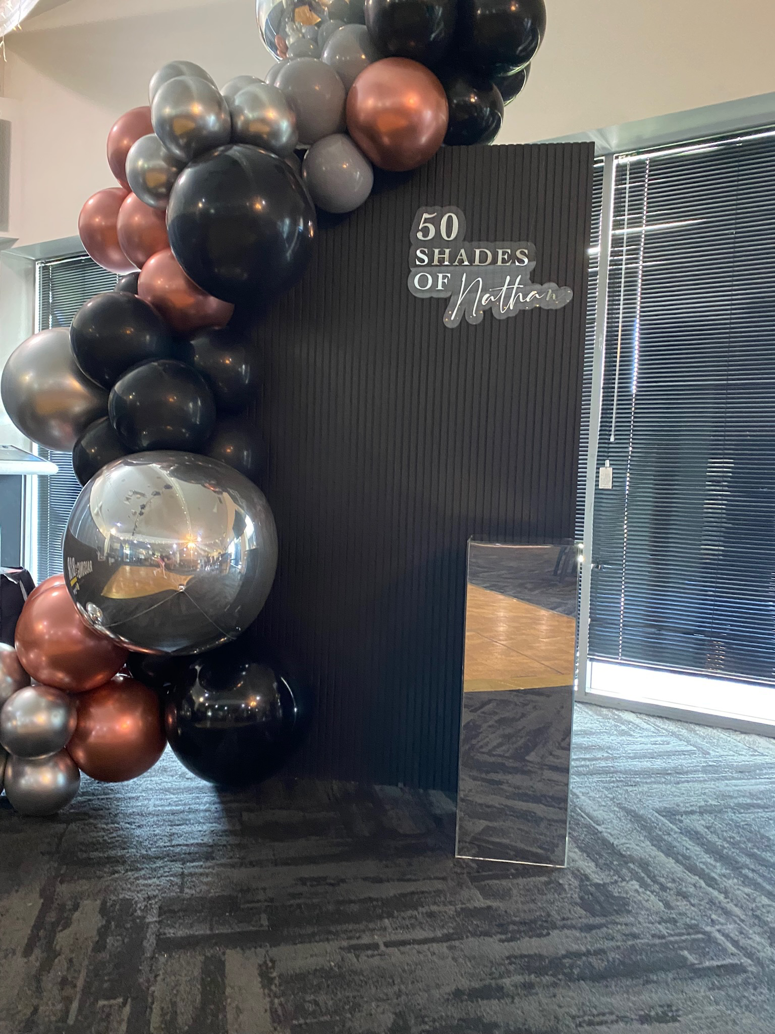 Black photo booth backdrop with a balloon arch in shades of black, gray, and metallic rose, and a sign that reads '50 Shades of Nathan'.