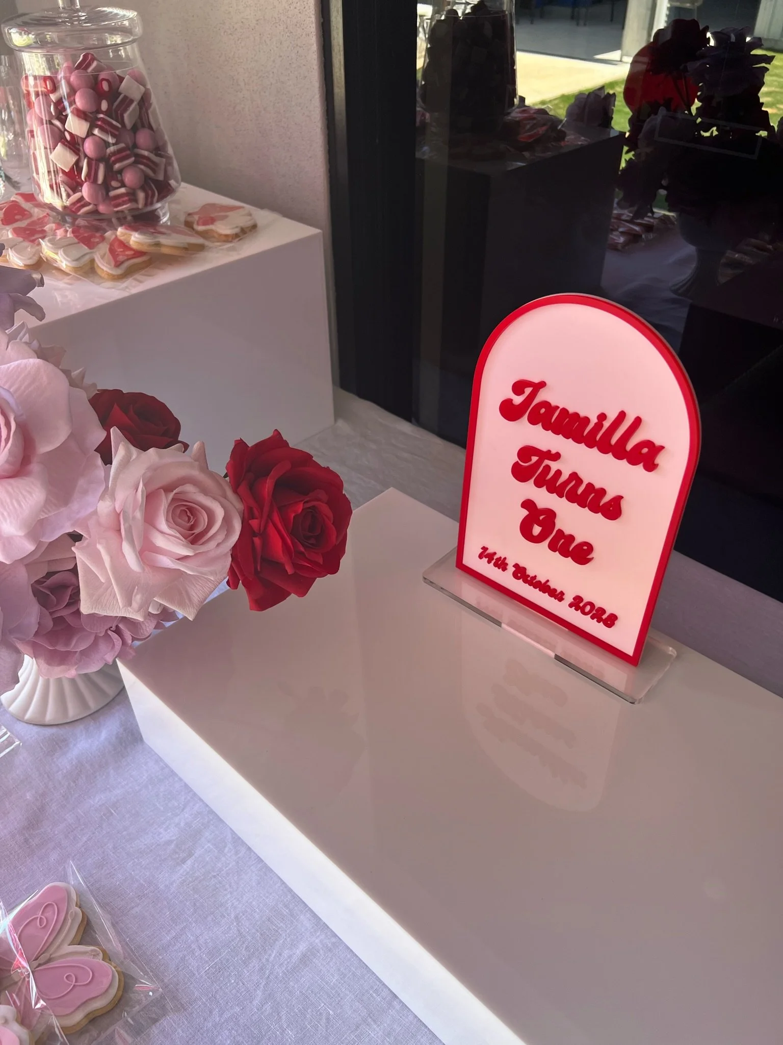 Table display for a birthday celebration with pink and red roses, a sign reading "Familia Turras One 14th October 2023," and pink heart-shaped cookies in the foreground.