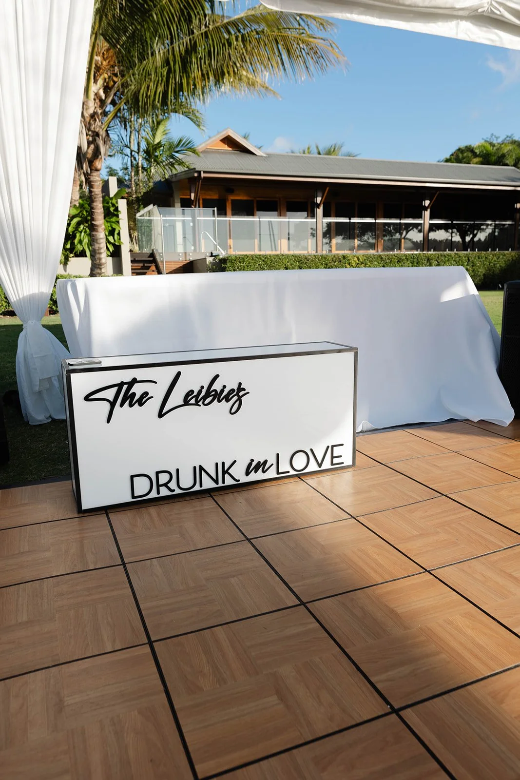 A white illuminated sign with black cursive and capital letters reads 'The Liebig' and 'DRUNK in LOVE' in front of a white table covered with a tablecloth on an outdoor wooden deck, with a background of palm trees, a house with a screened porch, and 