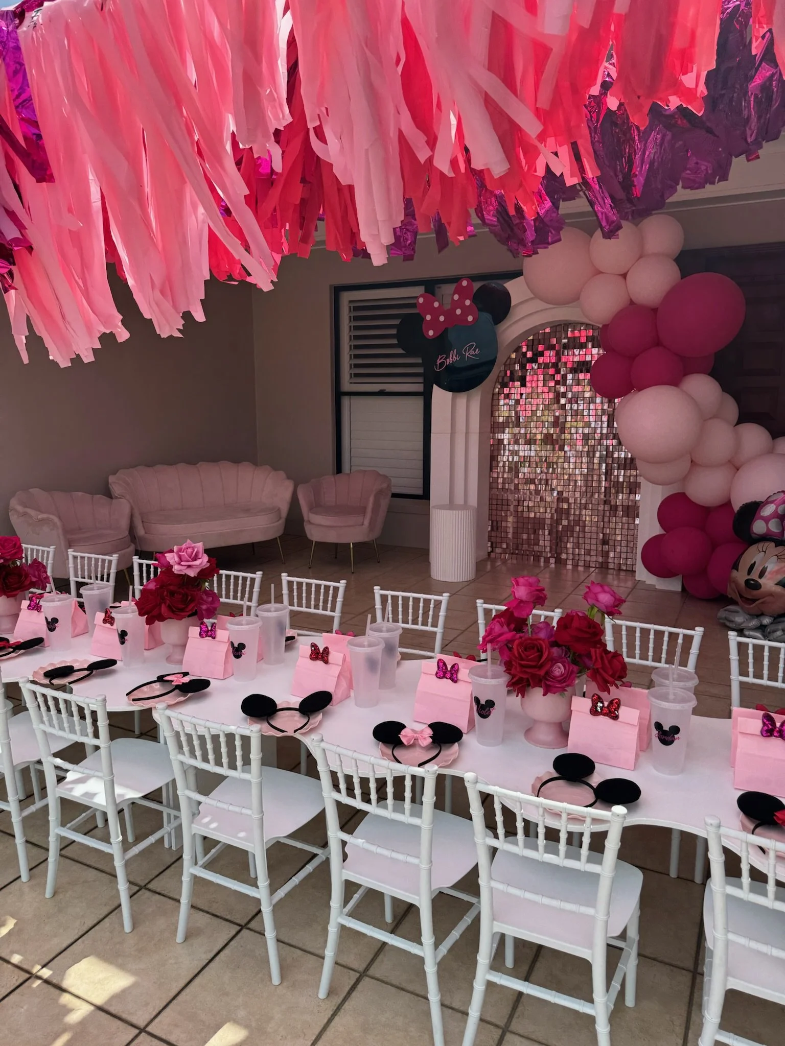 Decorated party room with pink and white balloons, Minnie Mouse themed accessories, pink flowers, and a long table with chairs, party favor boxes, and drinks, indicating a Minnie Mouse-themed birthday party for a young child.