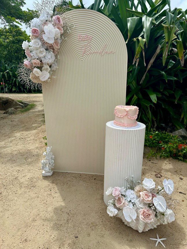Decorative baby shower setup with a cream-colored backdrop that says 'Baby Revelation,' a pink cake on a white pedestal, and floral arrangements with white and pink roses and white anthuriums, all outdoors on a sandy surface surrounded by greenery.