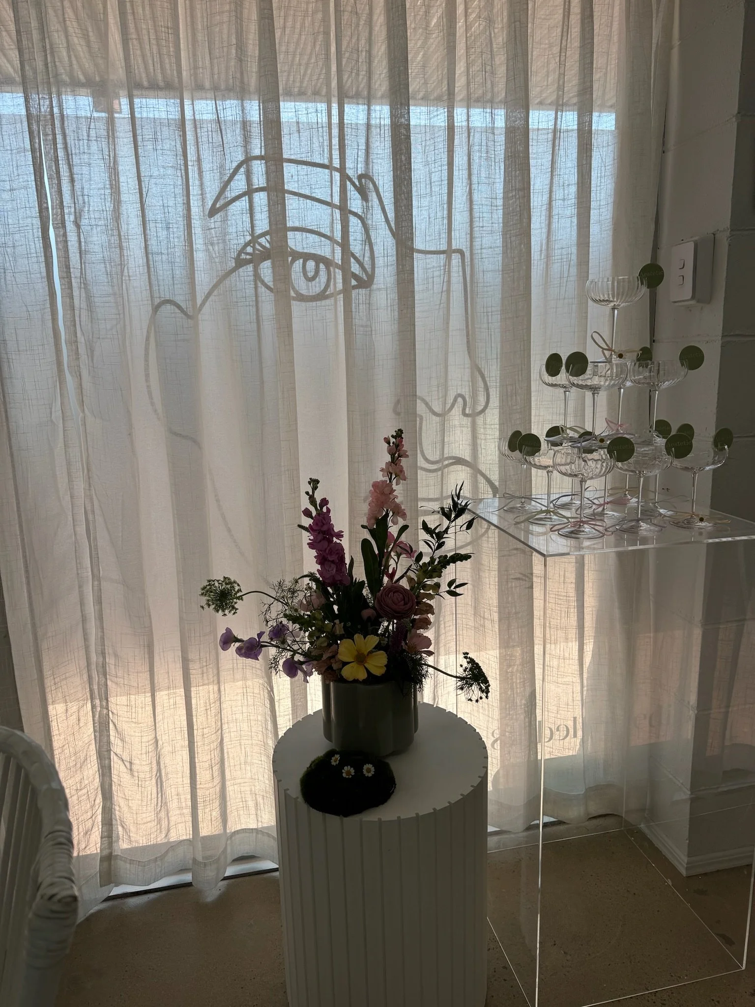 A vase with pink and purple flowers on a white, ribbed pedestal table in front of a sheer curtain with abstract line art of a face, and a champagne glass display on the right.