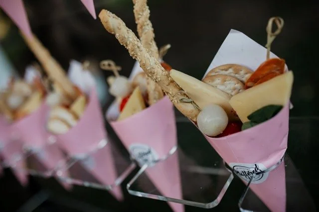 A gourmet cone with cheese, cherry tomato, breadstick, and crackers, placed in a pink paper lining on a metal stand.