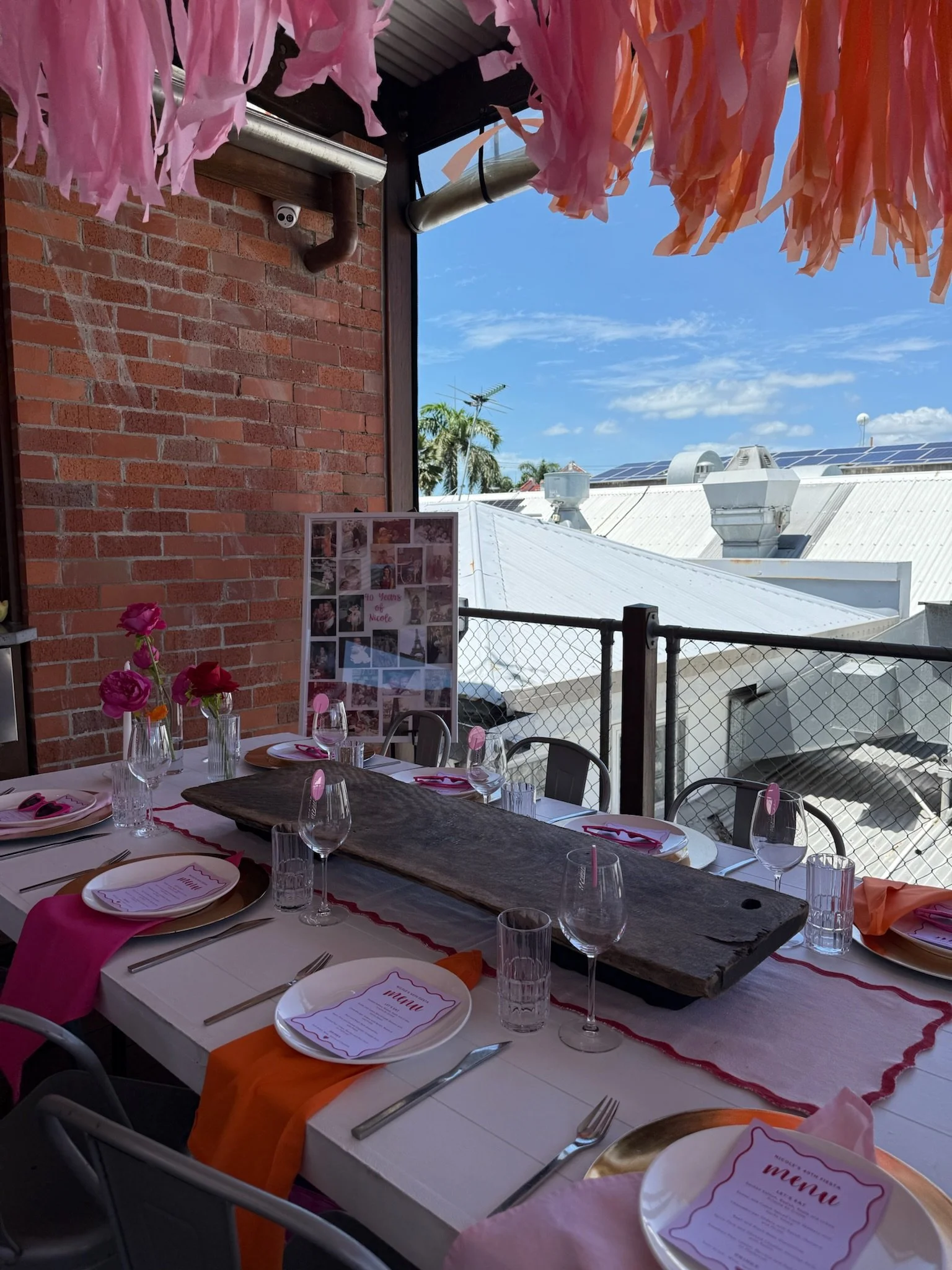 A decorated outdoor dining table with pink and orange accents, set for a meal with menus, glasses, cutlery, pink flowers in vases, and a centerpiece on a table runner, under pink tissue paper decorations with a brick wall and a building with solar pa