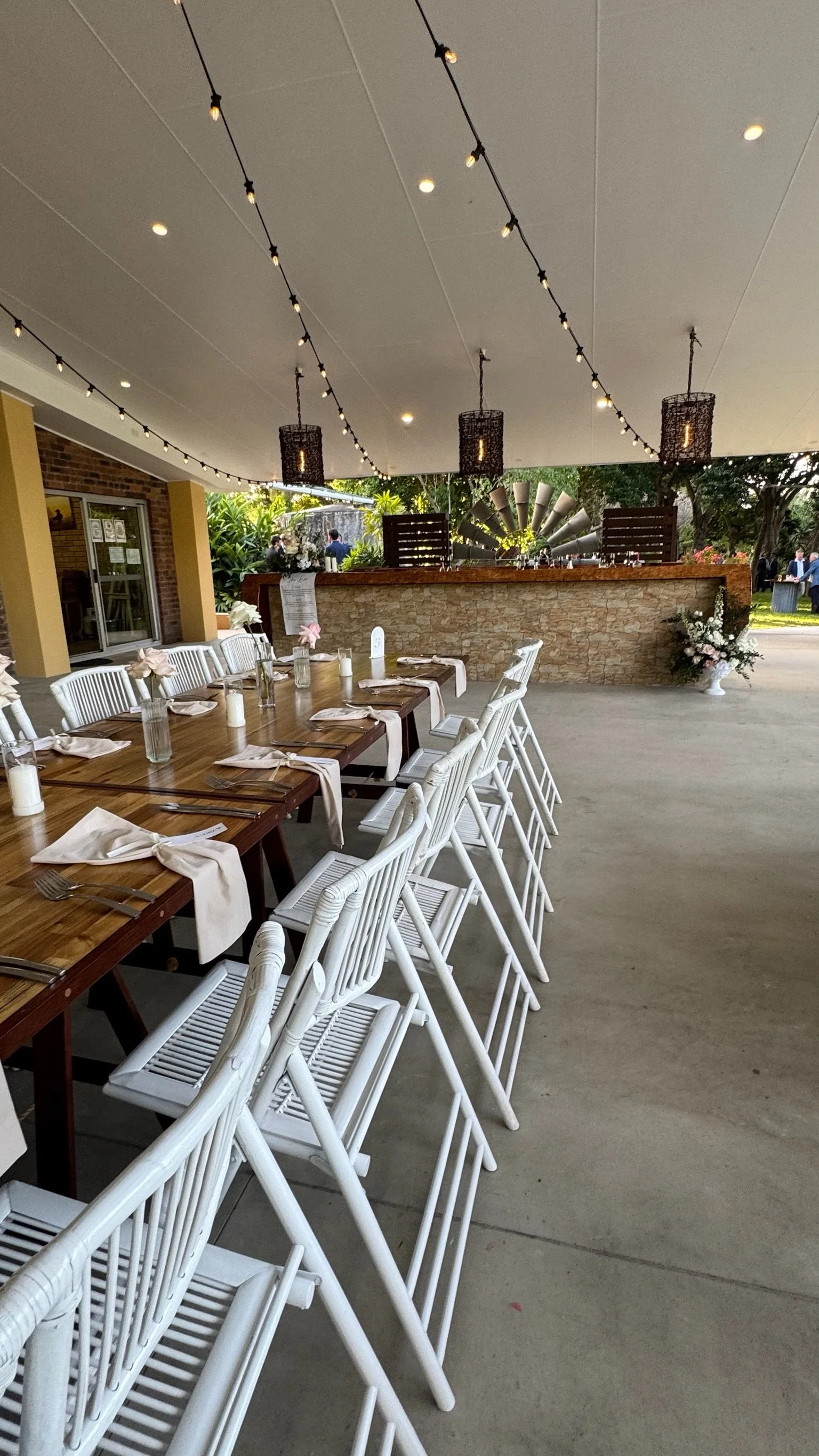 Outdoor event space with a long wooden table set with napkins, utensils, and candles, surrounded by white chairs. String lights hang overhead, and a bar with a stone facade is in the background, along with greenery and a few people.