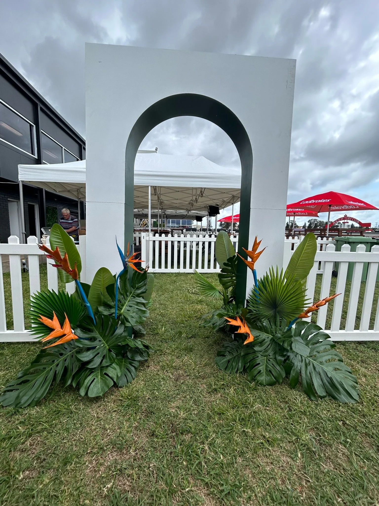 Decorative setup with tropical plants and orange bird of paradise flowers, arranged around a white archway on a grassy area, with overcast sky in the background.