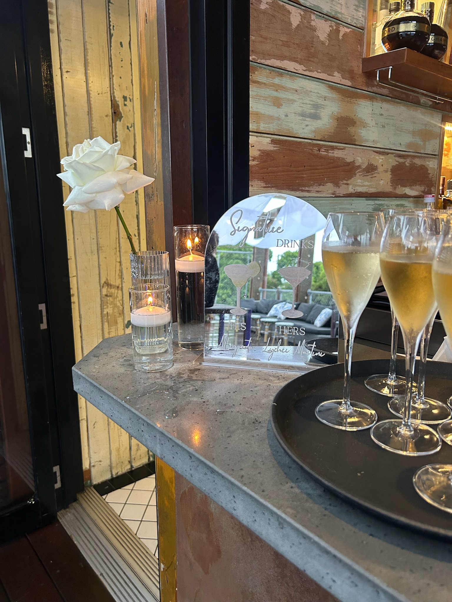 A decorative display with a white flower in a glass, two lit candles in tall holders, and four glasses of champagne on a tray, placed on a concrete countertop in a restaurant or bar setting with wooden wall panels.