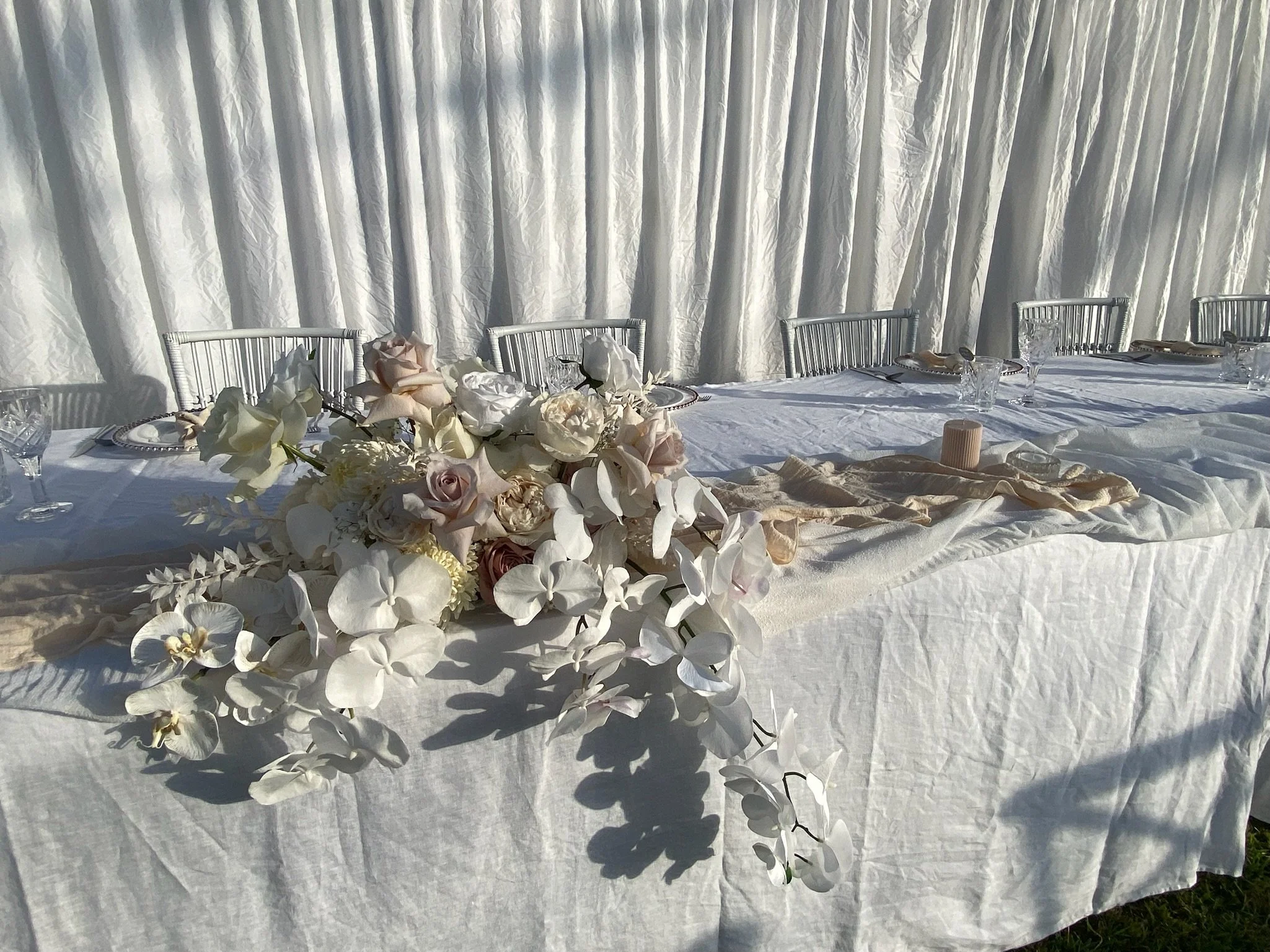 Elegant outdoor wedding or event table setup with white tablecloth, floral centerpiece of white and blush roses and orchids, glassware, plates, and gold napkins, set against white draped backdrop.