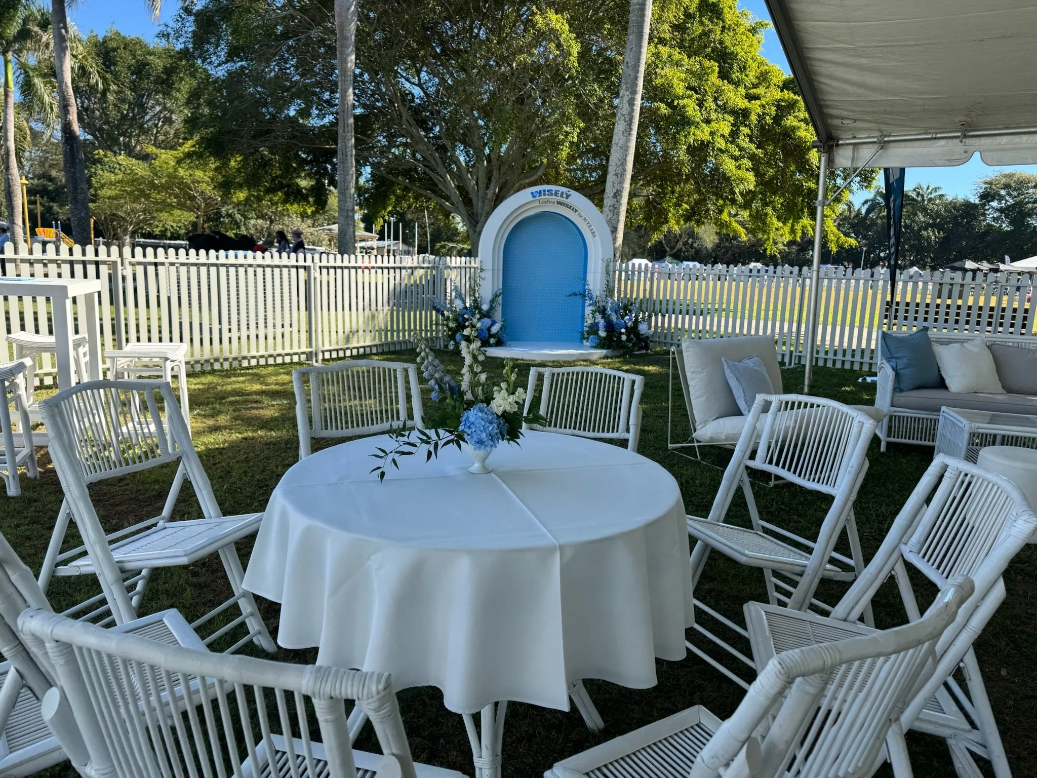 Outdoor wedding setup with white chairs around a round table with a floral centerpiece, a canopy, and a backdrop with flowers, garden, and a white picket fence.