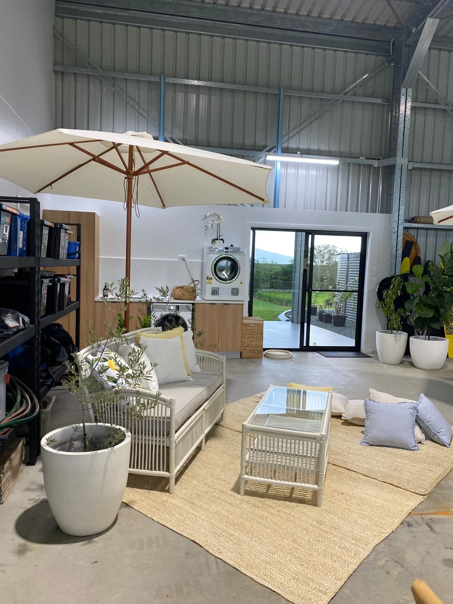 Indoor space with a glass coffee table, a white wicker sofa with pillows, large potted plants, an open door leading outside, and a laundry area with a washing machine and dryer.