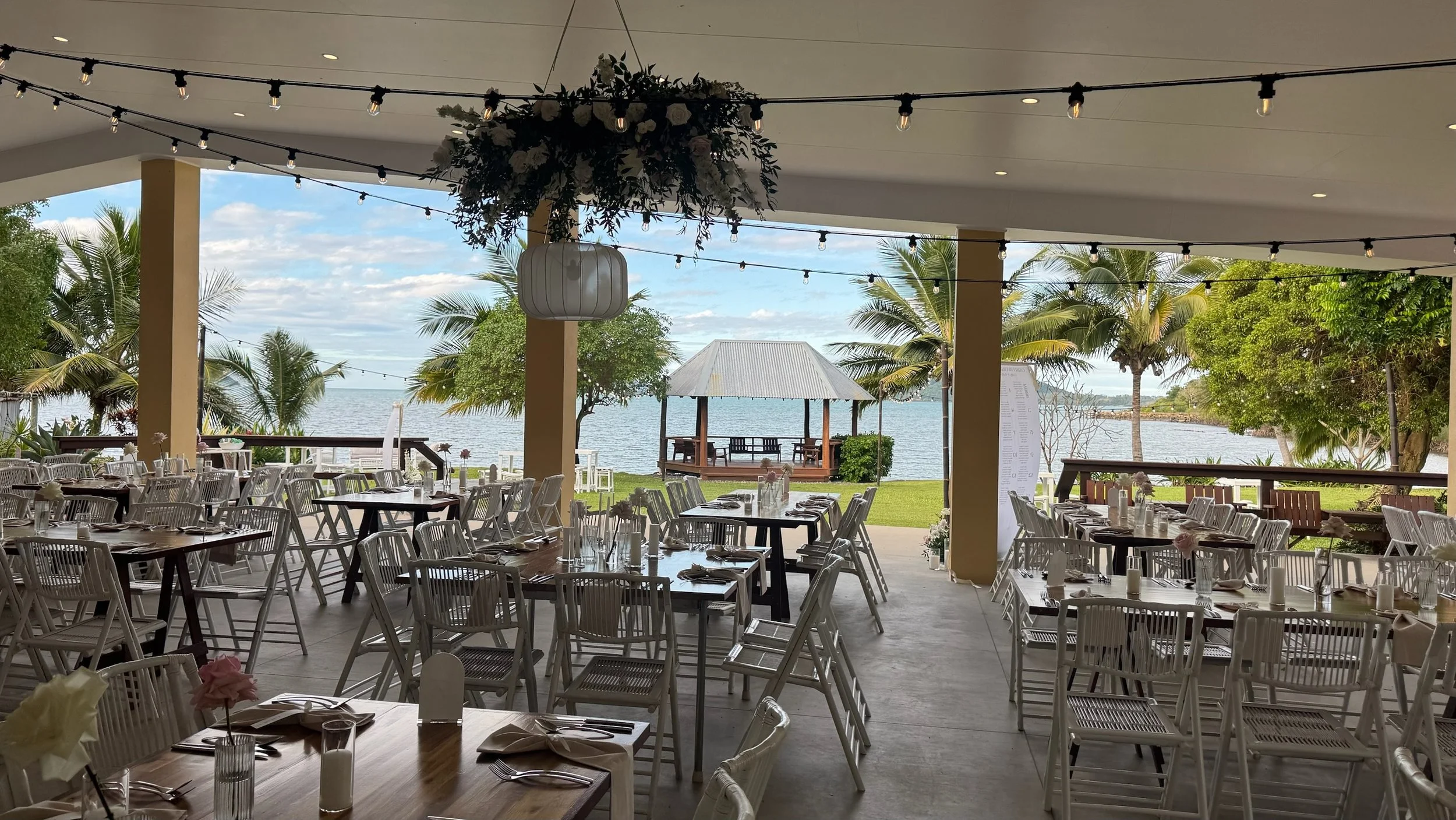 Seafood restaurant with outdoor dining area overlooking a body of water, decorated with string lights and floral centerpieces, with a gazebo and palm trees in the background.
