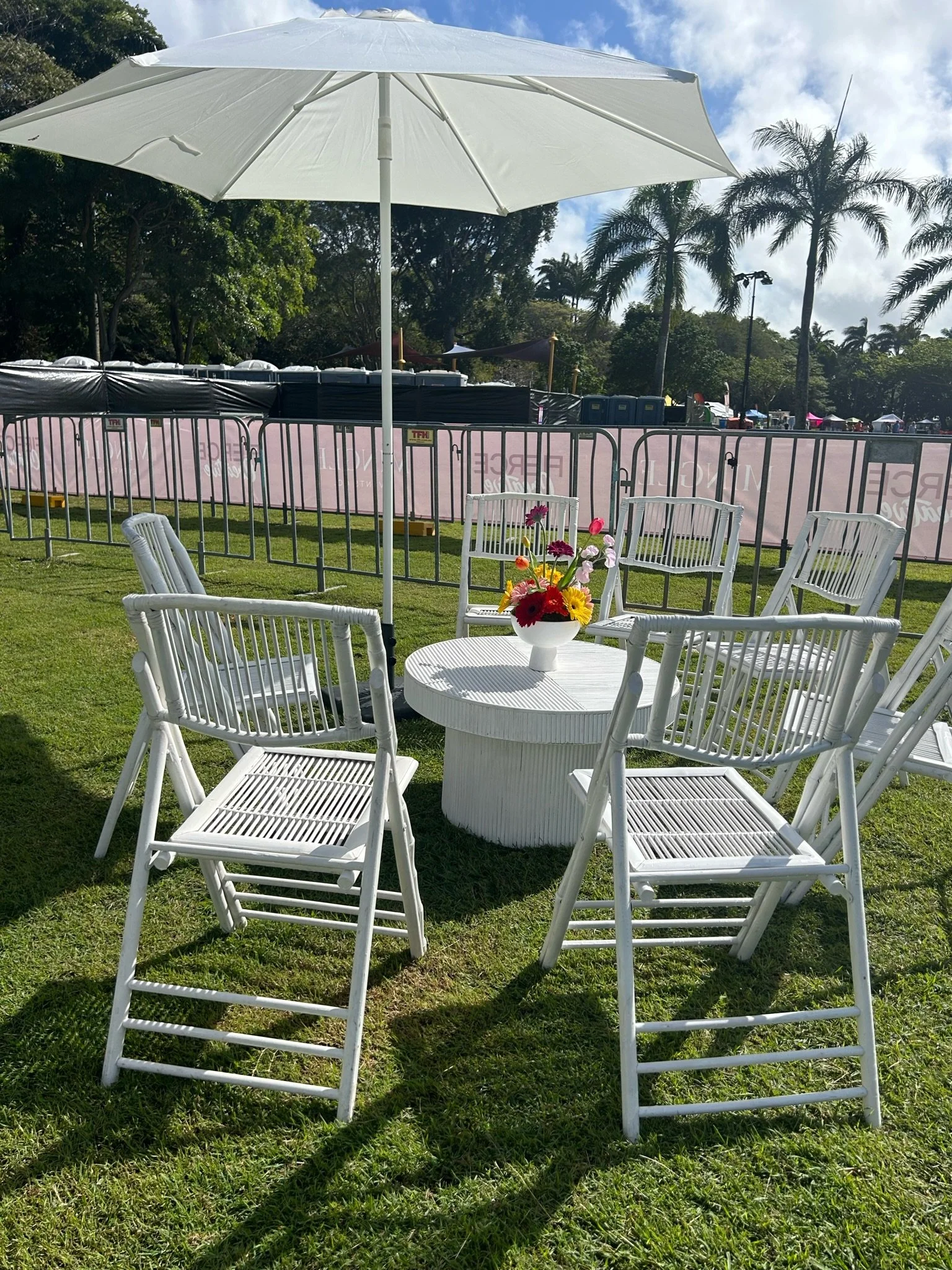 White patio table with a flower arrangement and six white chairs under a large white umbrella on a grassy lawn, with a pink barrier and palm trees in the background, under a partly cloudy sky.