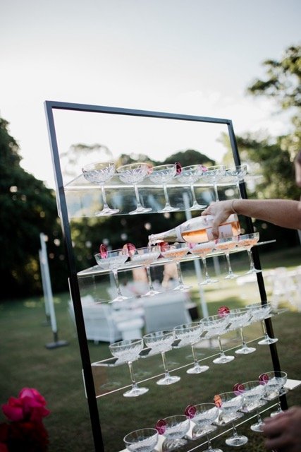 A person is filling champagne glasses on a tiered glass display at an outdoor event, with trees and a grassy area in the background.