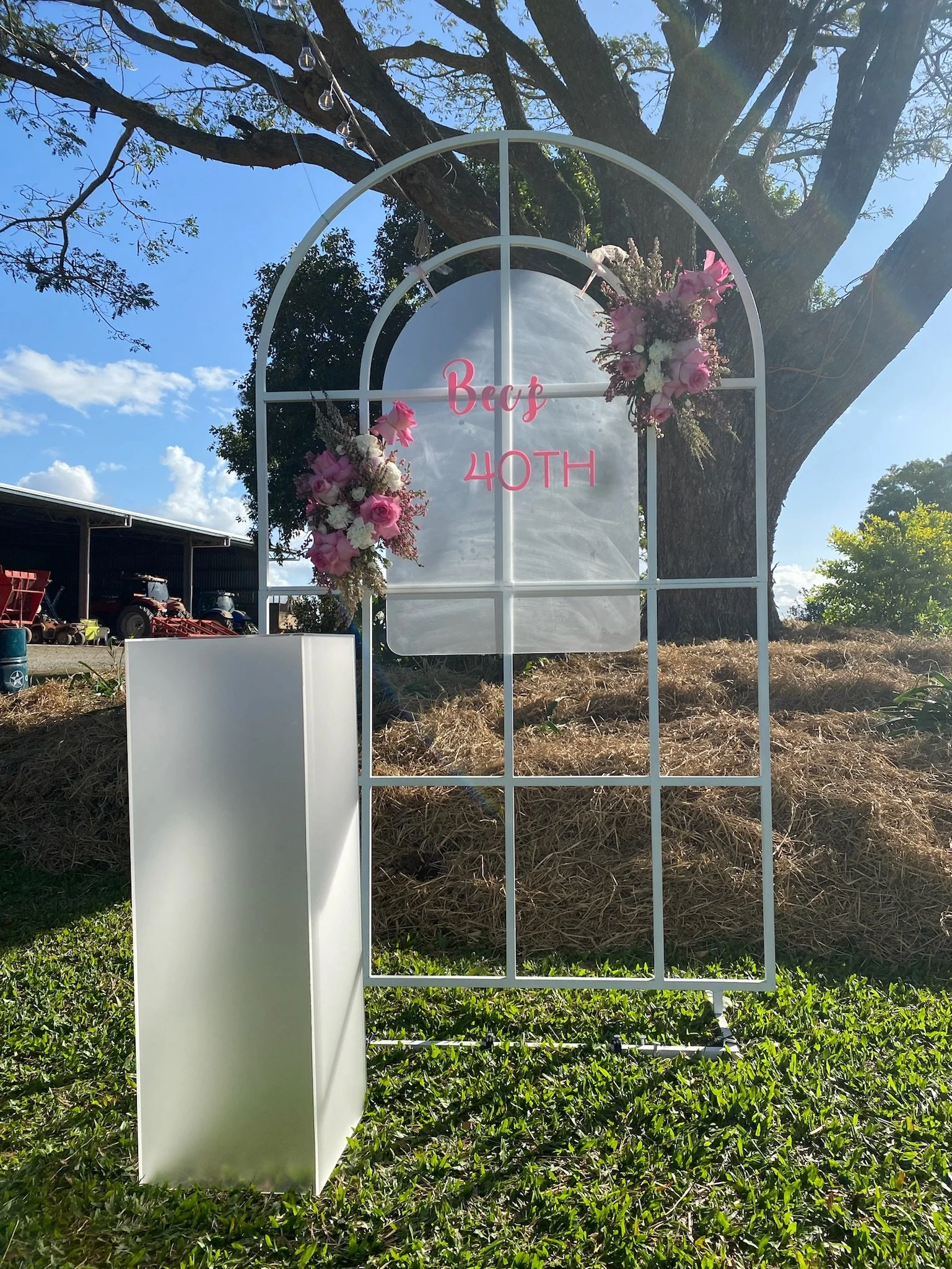 White garden arch decorated with pink and white flowers, with a sign referencing a 40th birthday celebration. The sign reads 'Bae 40TH' in pink lettering. The arch is set outdoors under a large tree on a grassy area, with a clear blue sky and some fa