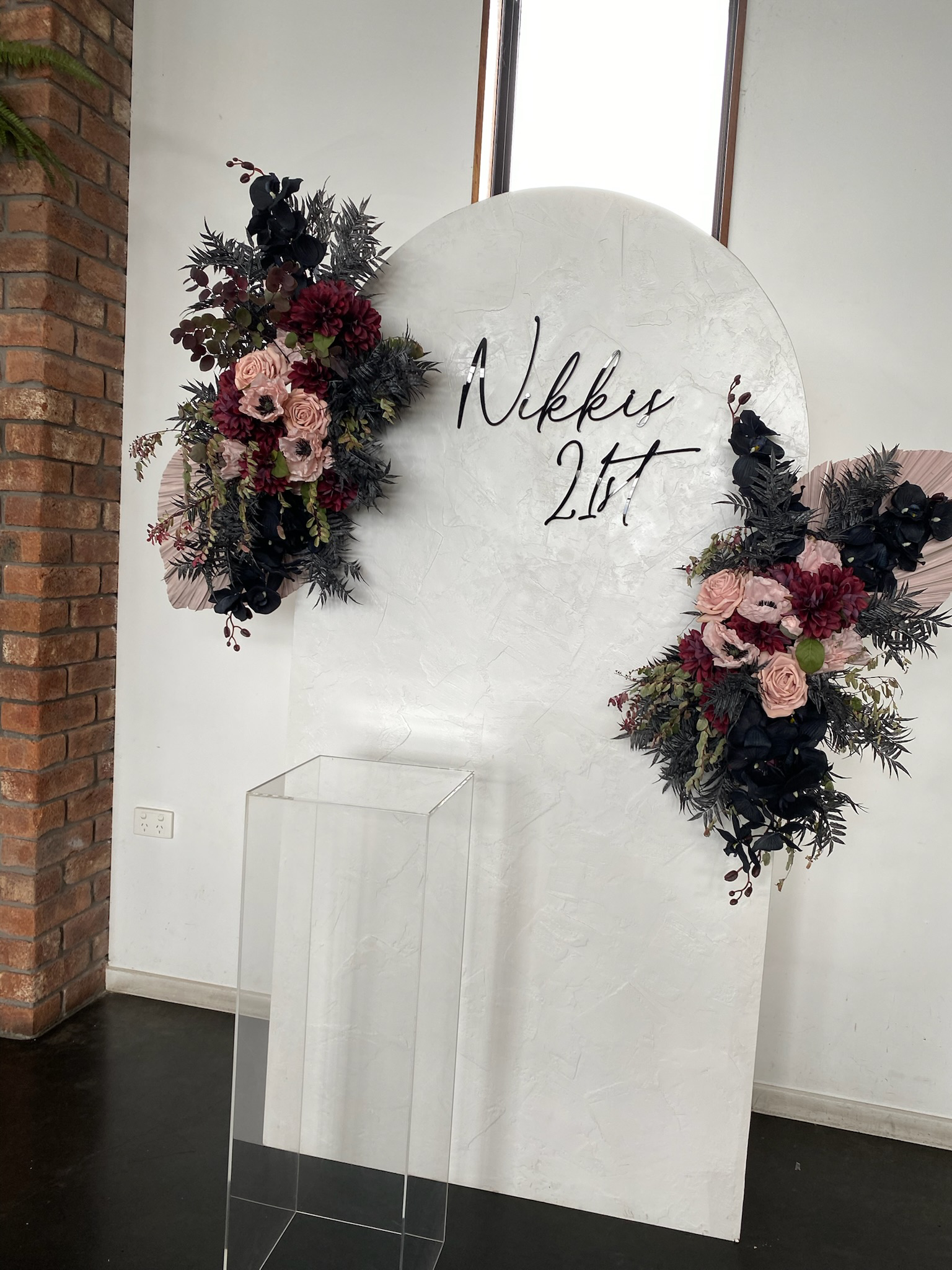 A white decorative panel with the text 'Nikkis 21st' in black cursive, adorned with floral arrangements of pink roses, dark red and black flowers, and greenery on the left and right sides.