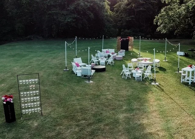 Outdoor event setup on a grassy field with white tables, chairs, cushions, and string lights, surrounded by trees.