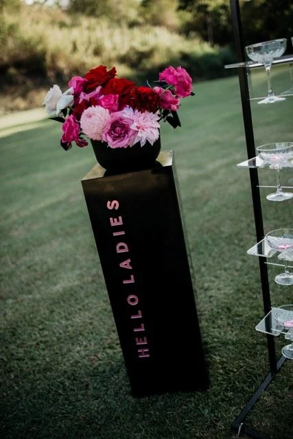 A floral arrangement in a black vase on a pedestal with pink and red flowers, next to a champagne glass display on a golf course.