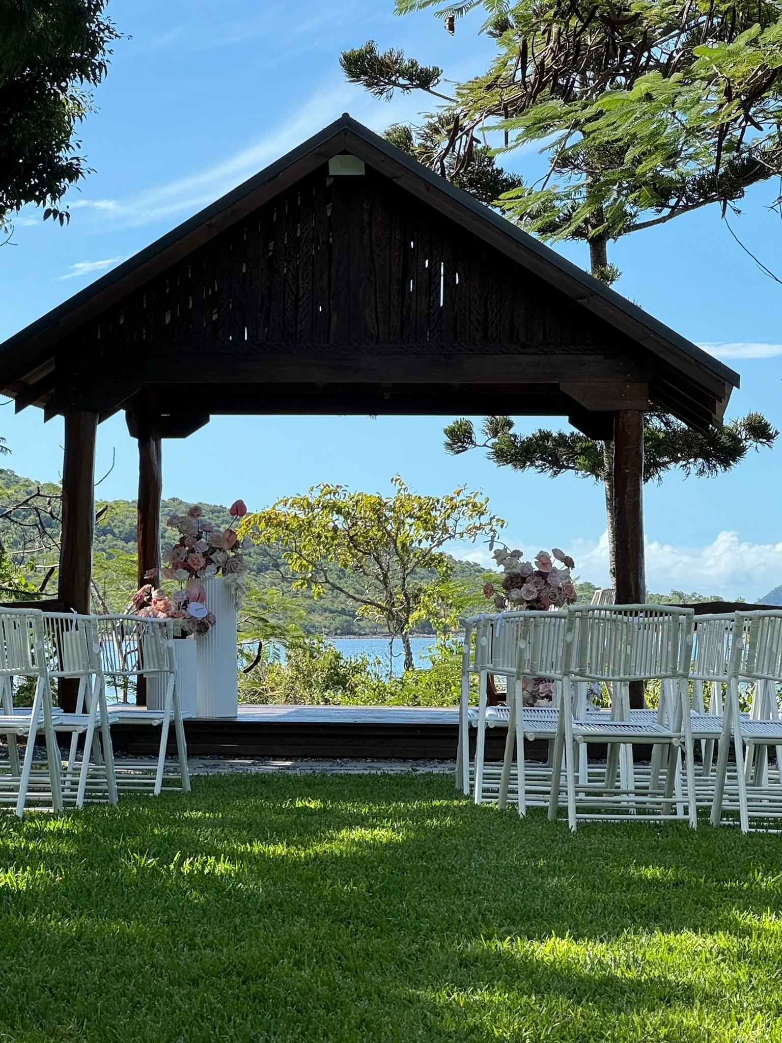 Outdoor wedding setup with white chairs arranged on a green lawn, decorated with pink and white flowers, under a wooden pavilion overlooking a lake with trees and hills in the background.
