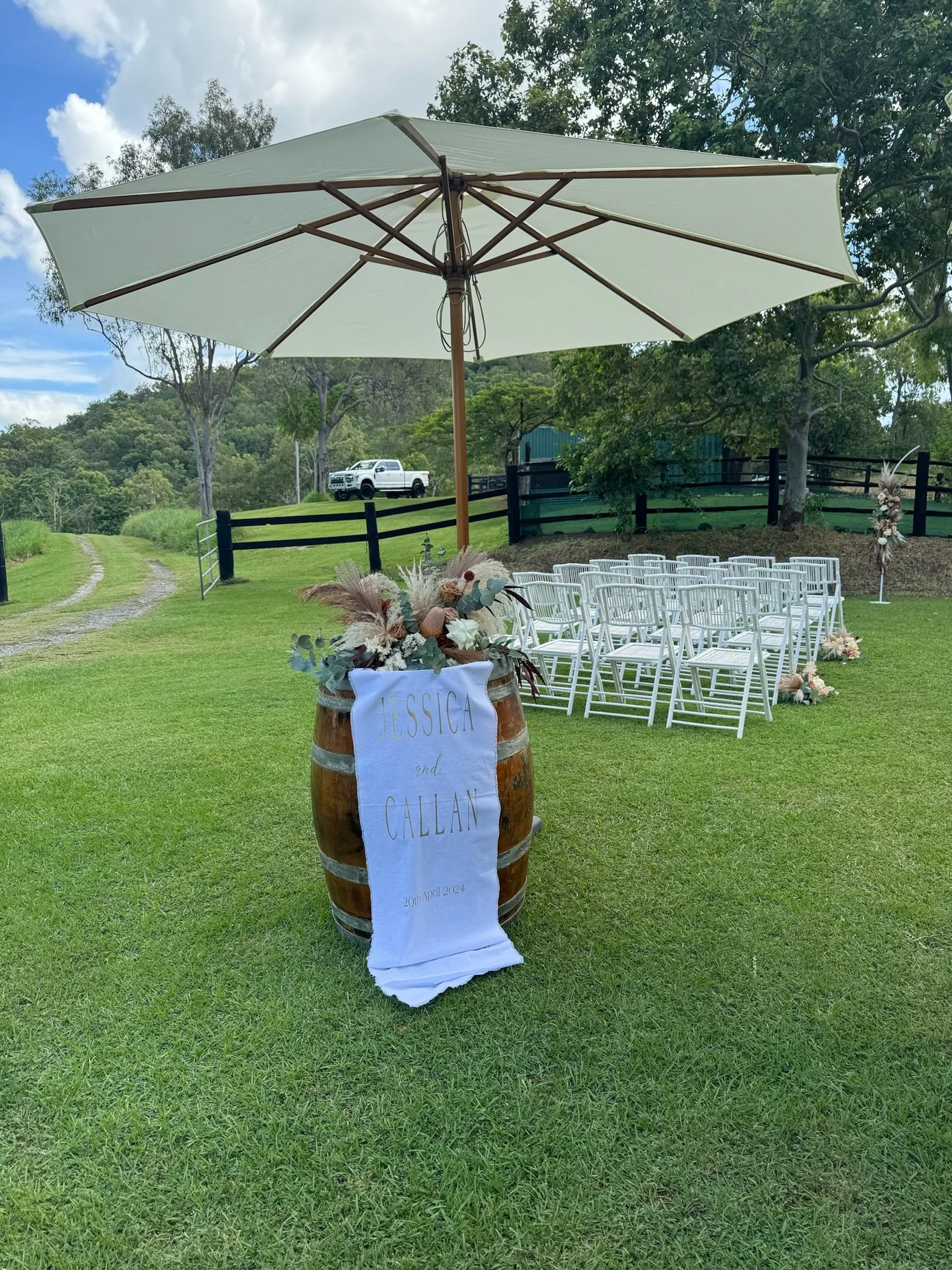 Outdoor wedding setup on a grassy field with white chairs arranged for guests, a wooden barrel decorated with flowers and a banner with names and date, a large white patio umbrella providing shade, trees in the background, and a white pickup truck pa