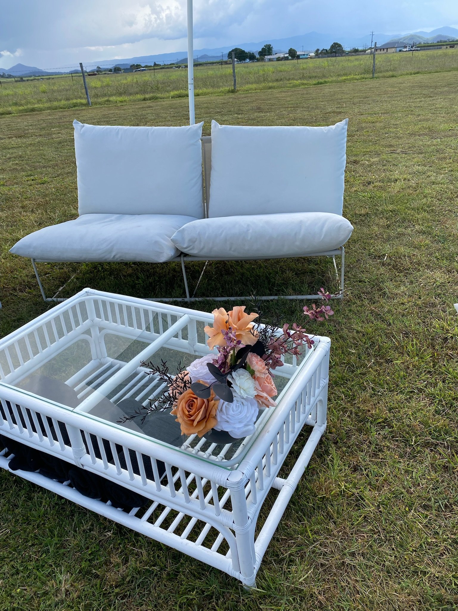 White outdoor lounge chair with two cushions behind a white wicker table with a floral arrangement on top, set on a grassy field under a cloudy sky.