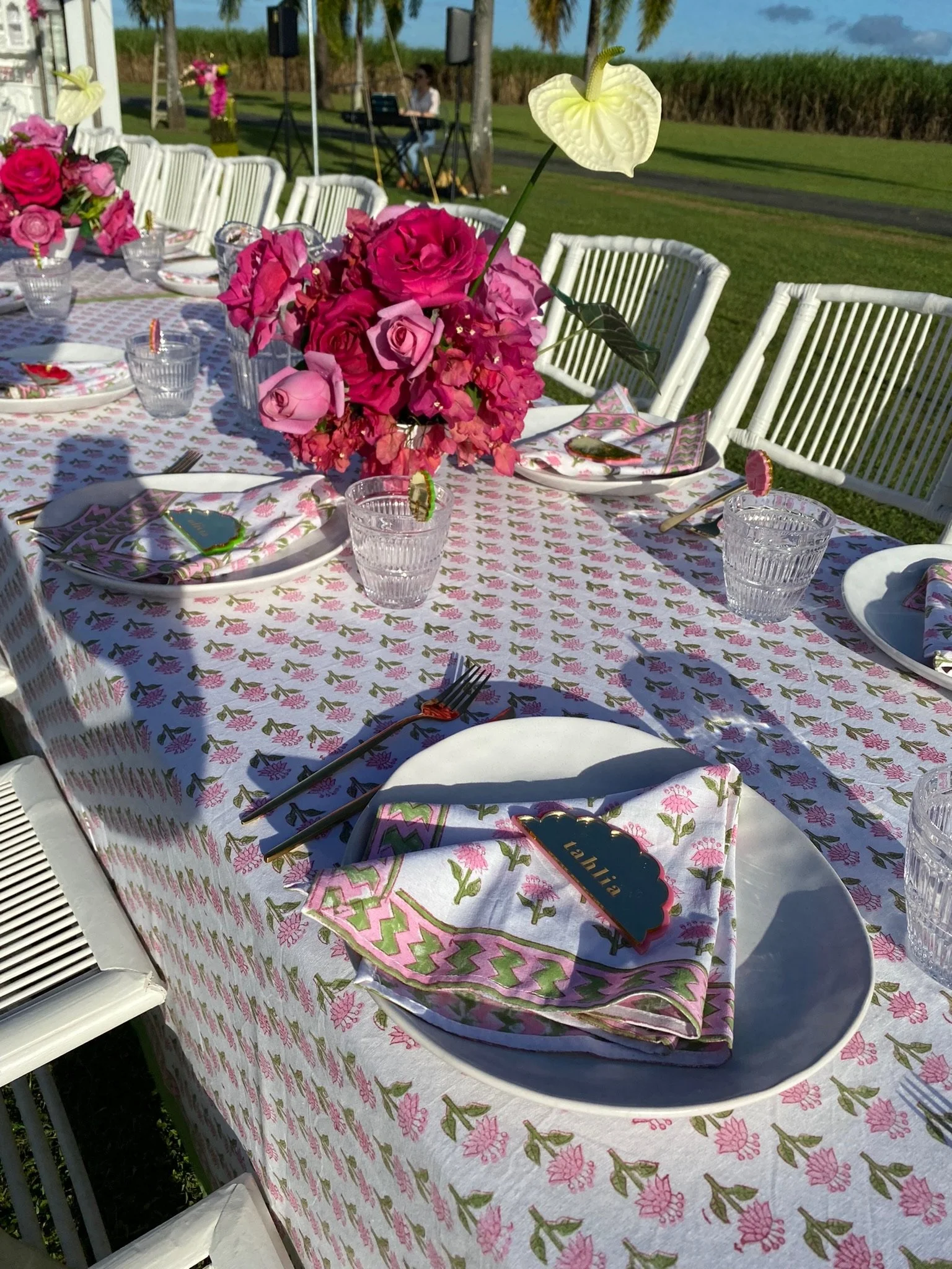 Outdoor table set for a celebration with pink floral centerpiece, white plates, napkins, and clear glasses on a pink and white floral tablecloth. There are name tags on the napkins, and in the background, there is a grassy area with a person at a key