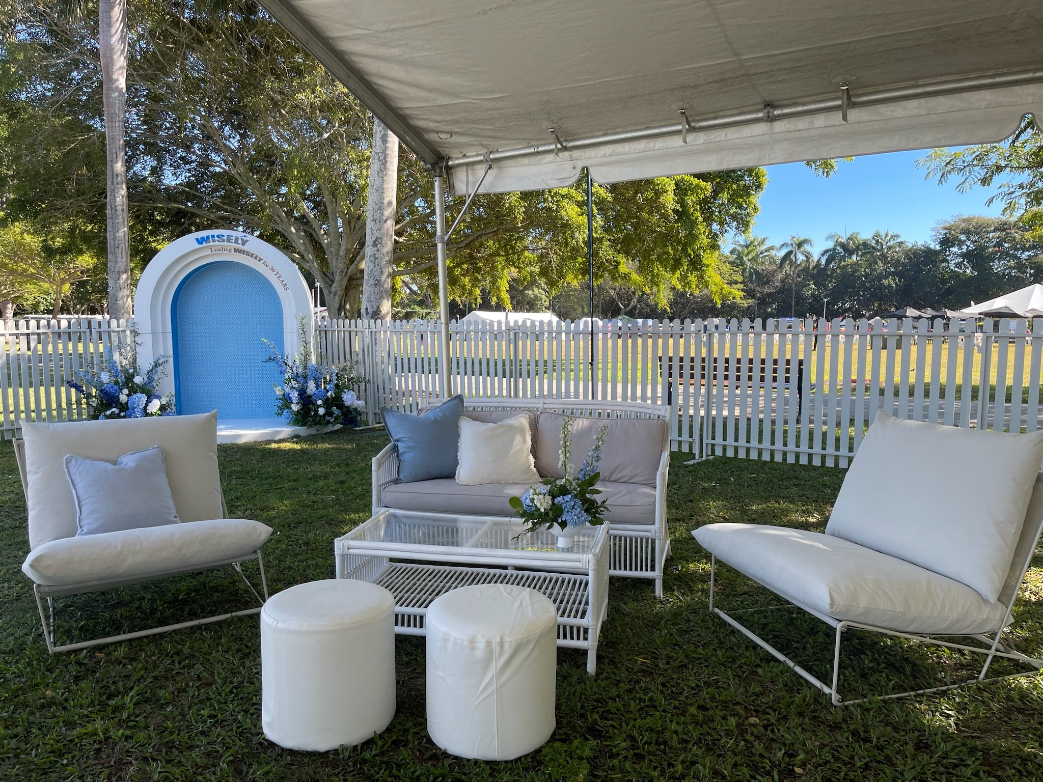 Outdoor event setup with white cushioned seating, a small glass table with a flower arrangement, and a white decorative fountain with blue flowers in a grassy area enclosed by a white picket fence, under a canopy with trees and a sunny sky in the bac