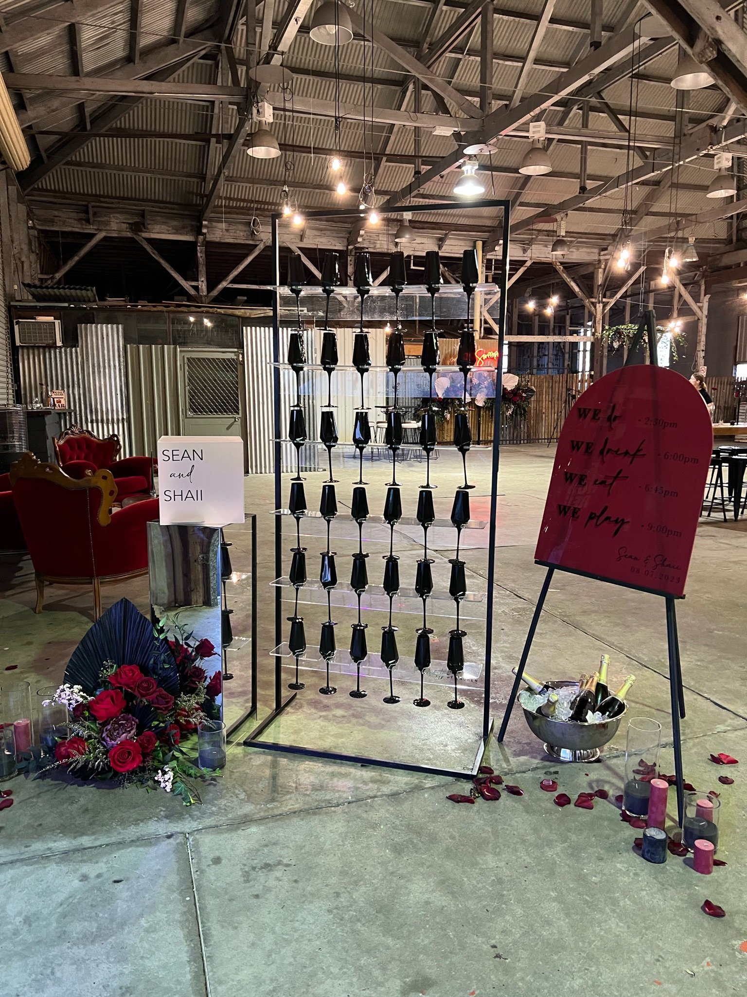 Decorative setup for a wedding reception featuring a black bottle display, a large pink sign with event times, a floral arrangement, candles, and a vintage armchair in an industrial-style venue with exposed beams and string lights.