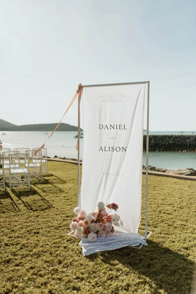Wedding or welcoming sign with names Daniel and Alison near a beach, decorated with flowers, set up on grass with chairs and ocean in the background.