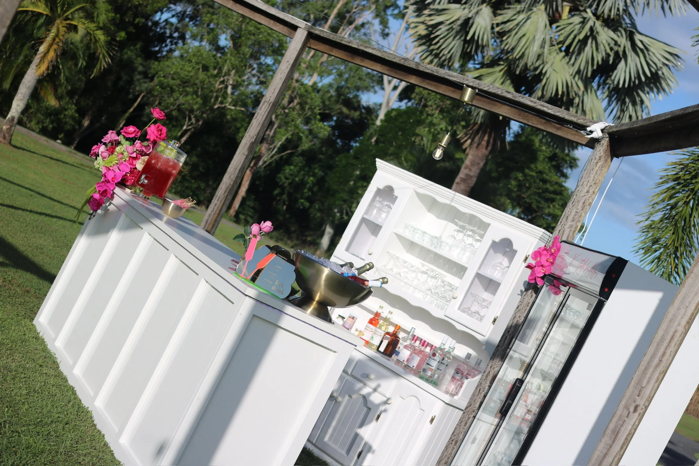 Outdoor bar setup with white cabinetry, glassware, bottles, and pink flowers under a wooden frame in a tropical garden.