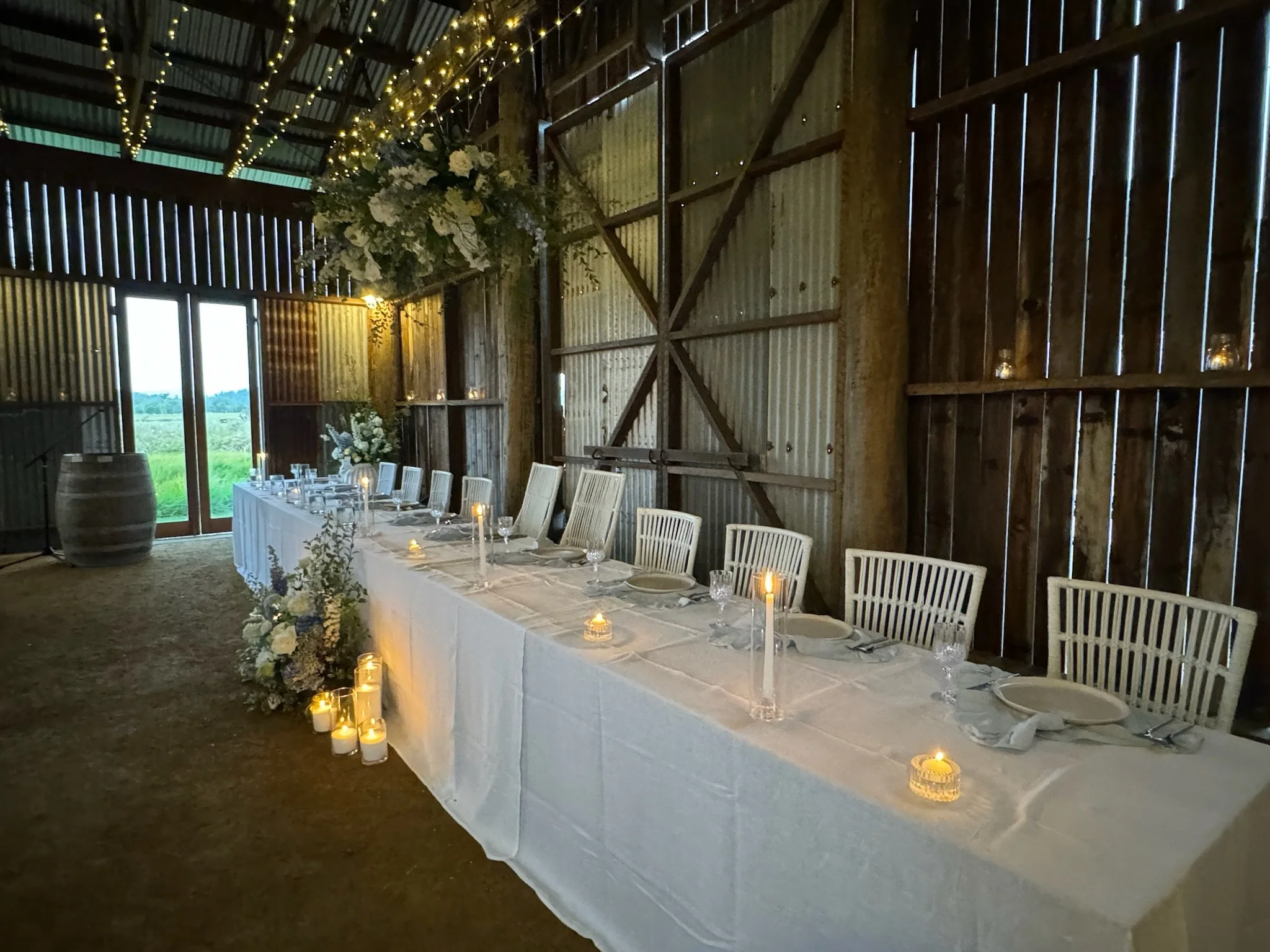 Long rectangular dining table with white tablecloth, place settings, and glassware, arranged for a celebration or wedding in a rustic barn with candles and floral decorations, ambient lighting, and wooden walls.