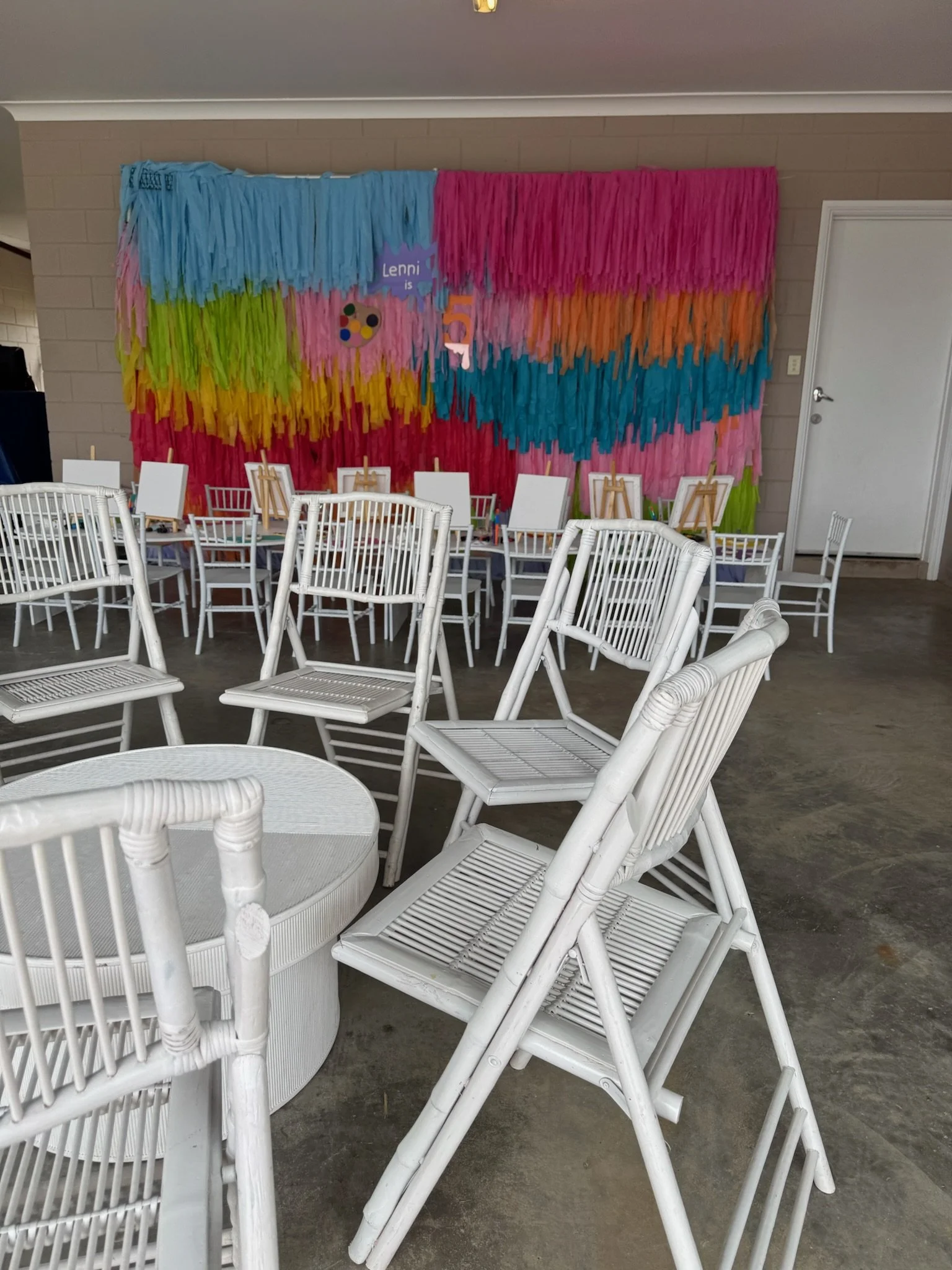 Colorful DIY backdrop with streamers in pink, blue, orange, and green for a party, with empty white chairs and small easels in a room.