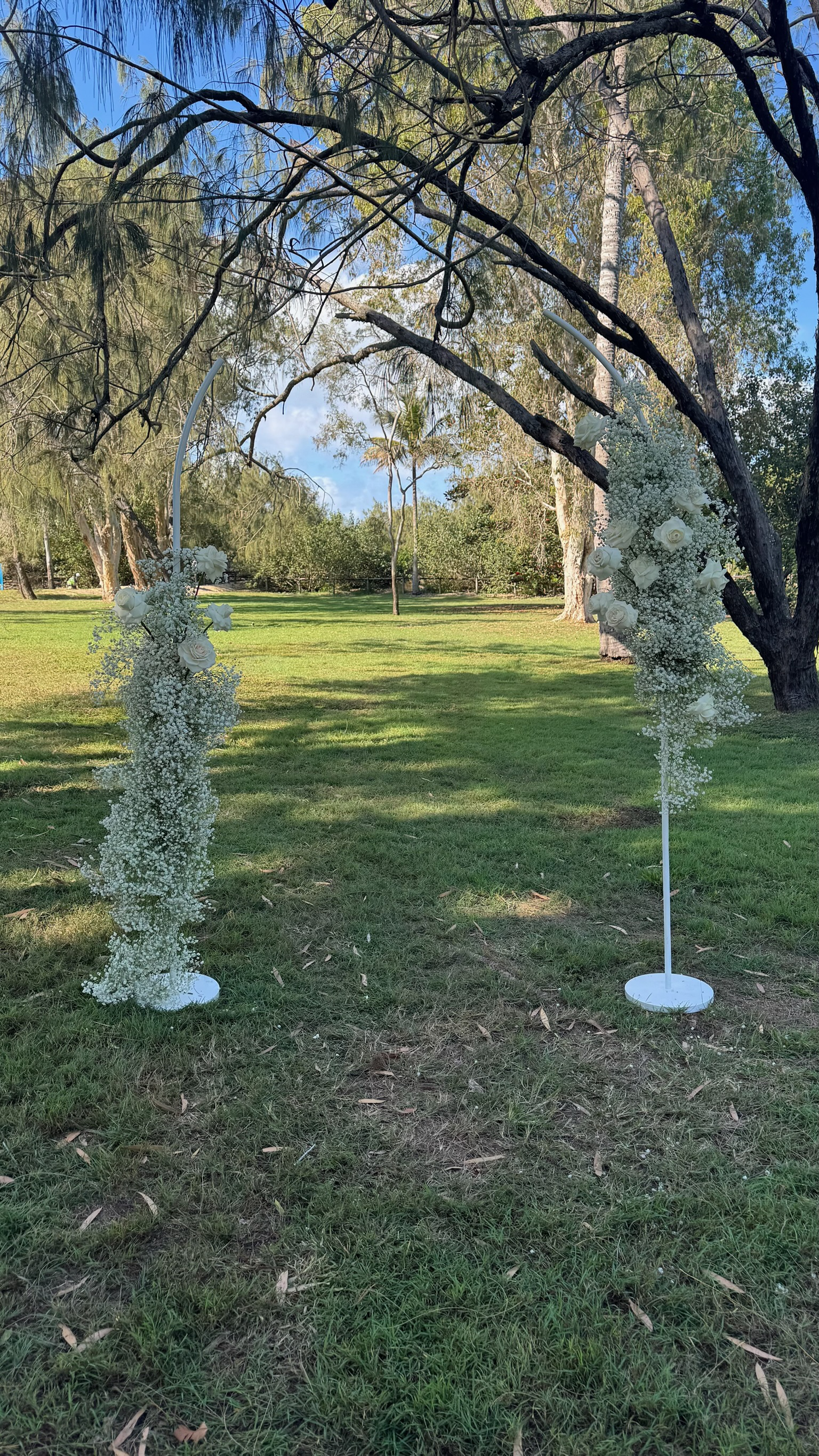 Decorative white floral arch with white roses and baby's breath, set up outdoors on grass under trees, likely for a wedding ceremony.
