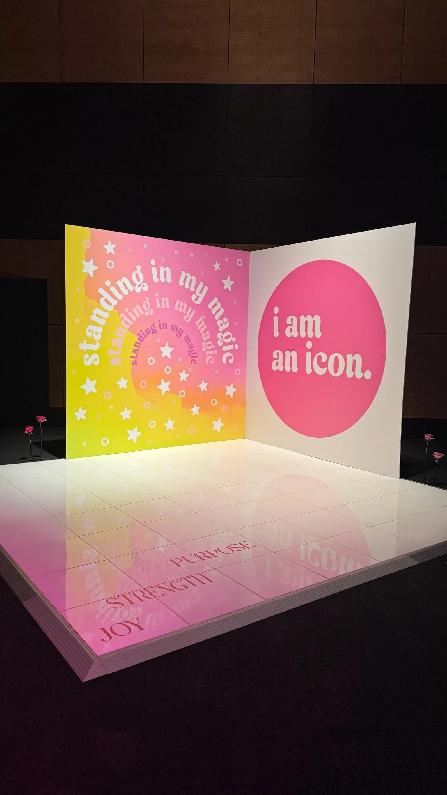 Colorful wall display with pink, yellow, and orange background, featuring text about standing in one's magic and being an icon, with words like 'strength,' 'joy,' 'purpose' reflected on the floor.