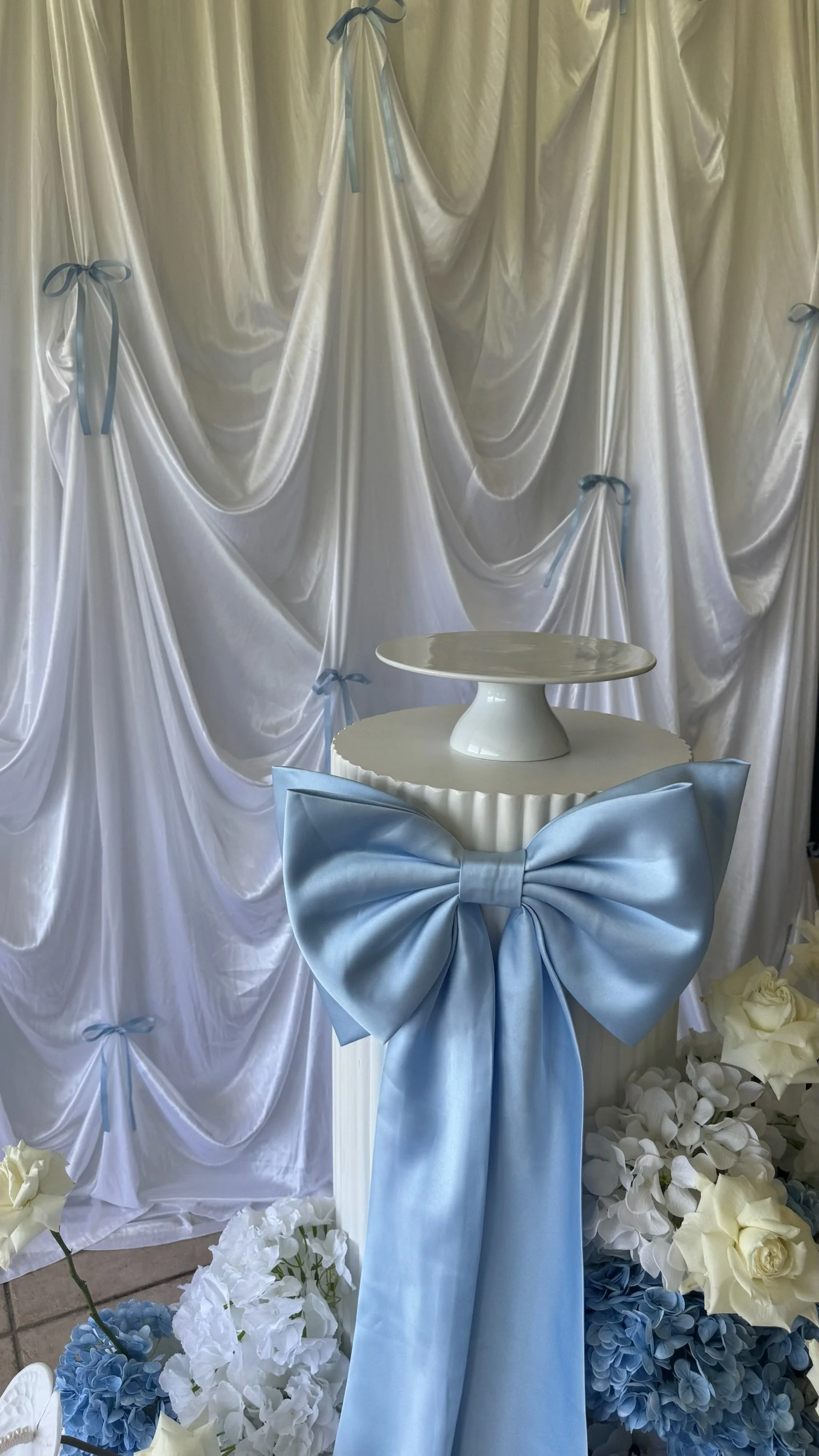 Decorative display with a white cake stand draped in a large light blue satin bow, set against white satin drapery with blue ribbons and surrounded by white and blue flowers.