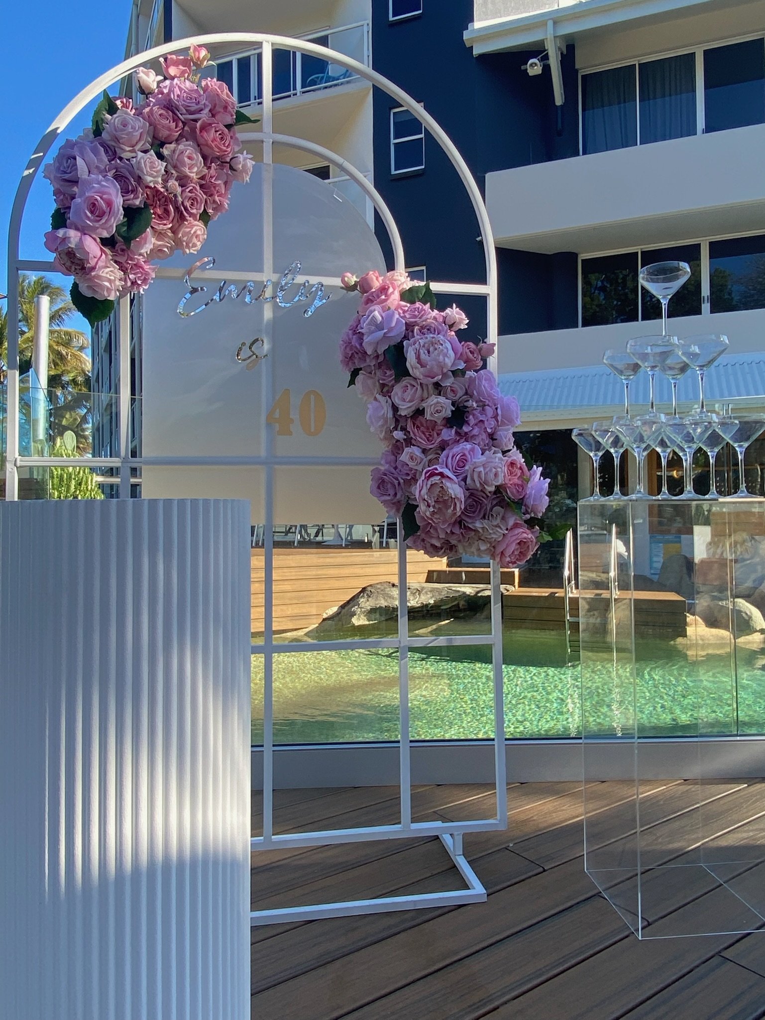 A decorated wedding or celebration arch with pink and purple roses, a sign reading 'Evely & 40', set outdoors next to a swimming pool with stacked glasses nearby, in front of a modern building with blue walls.