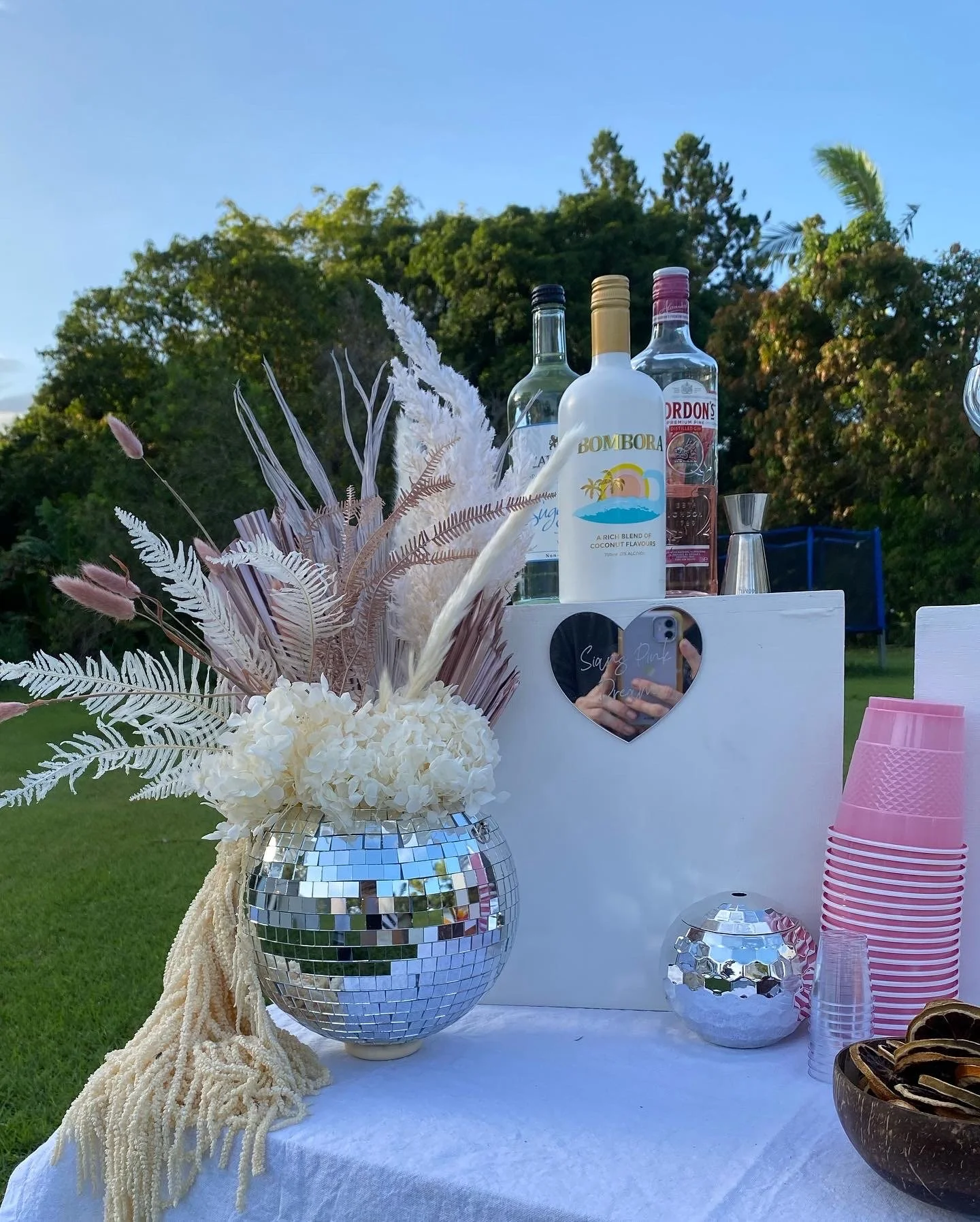 Outdoor party setup with a table holding a reflective mirror ball flower arrangement, bottles of alcohol, pink plastic cups, and a heart-shaped mirror with writing.
